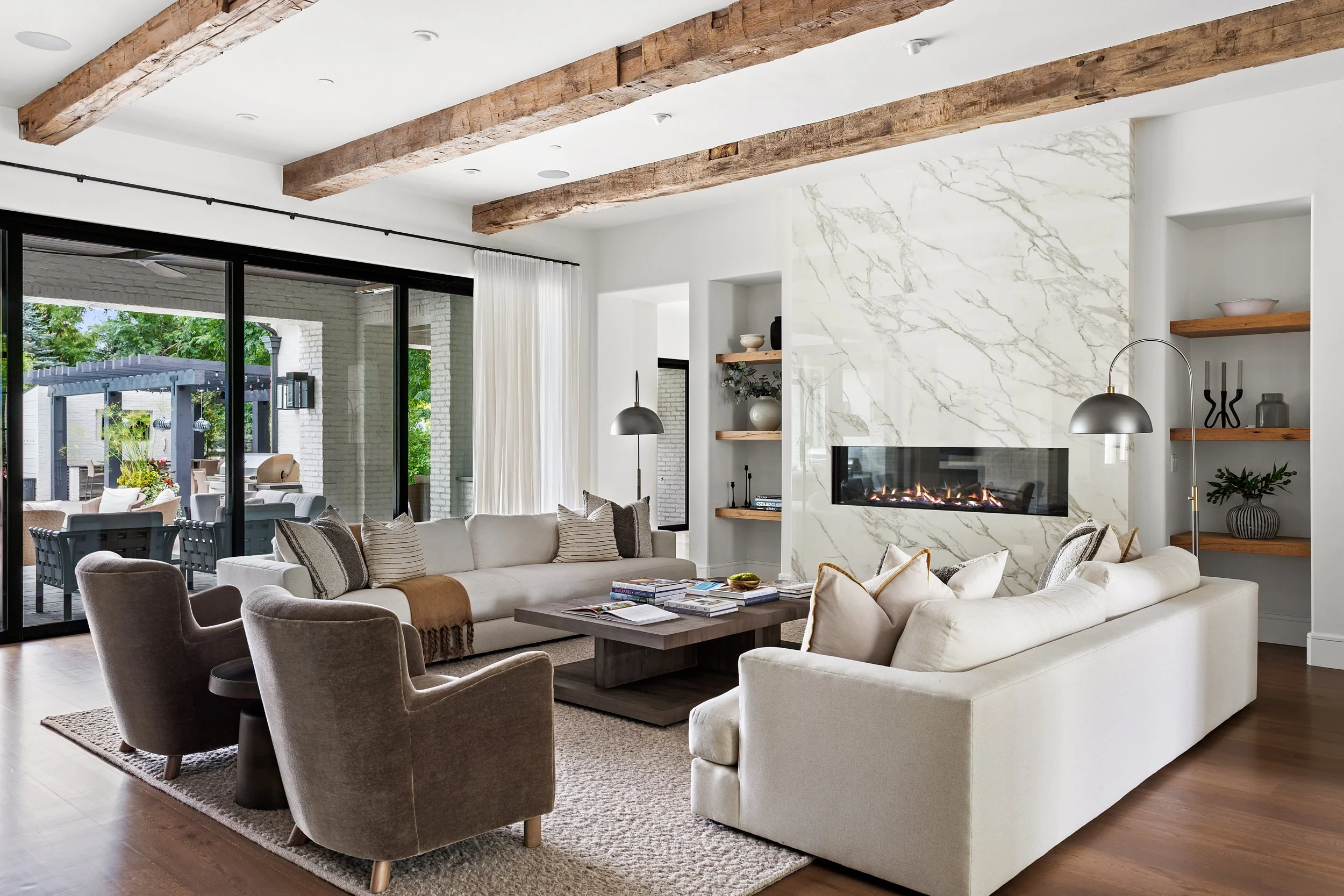 A spacious living room with white walls, a marble fireplace, wooden ceiling beams, comfortable sofas, and a large sliding glass door leading to an outdoor patio with greenery.