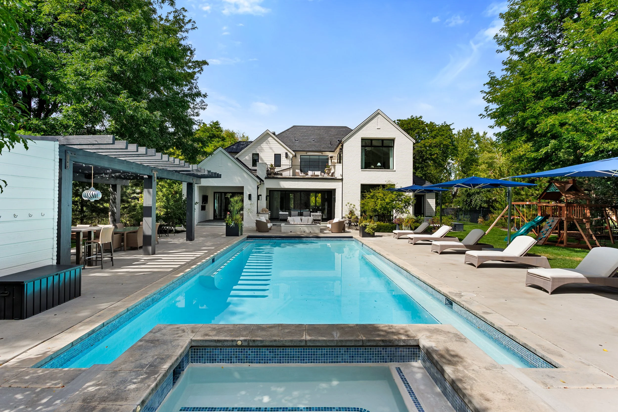 A backyard with a swimming pool, lounge chairs, and a house with a balcony, surrounded by green trees under a blue sky.