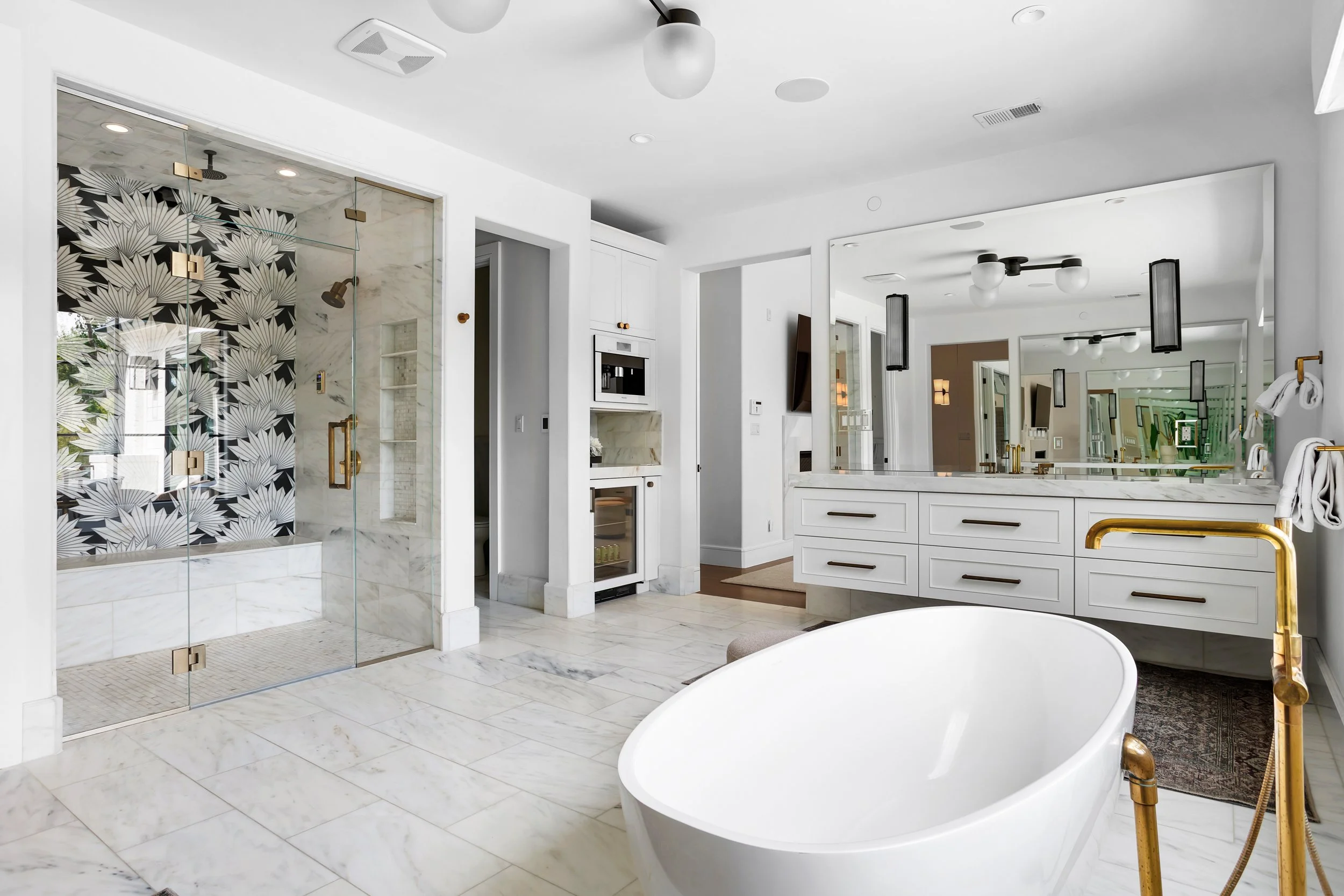 Modern bathroom with a freestanding bathtub, white marble floors, a glass shower enclosure with black and white patterned tile, a white vanity with gold fixtures, and large mirrors.