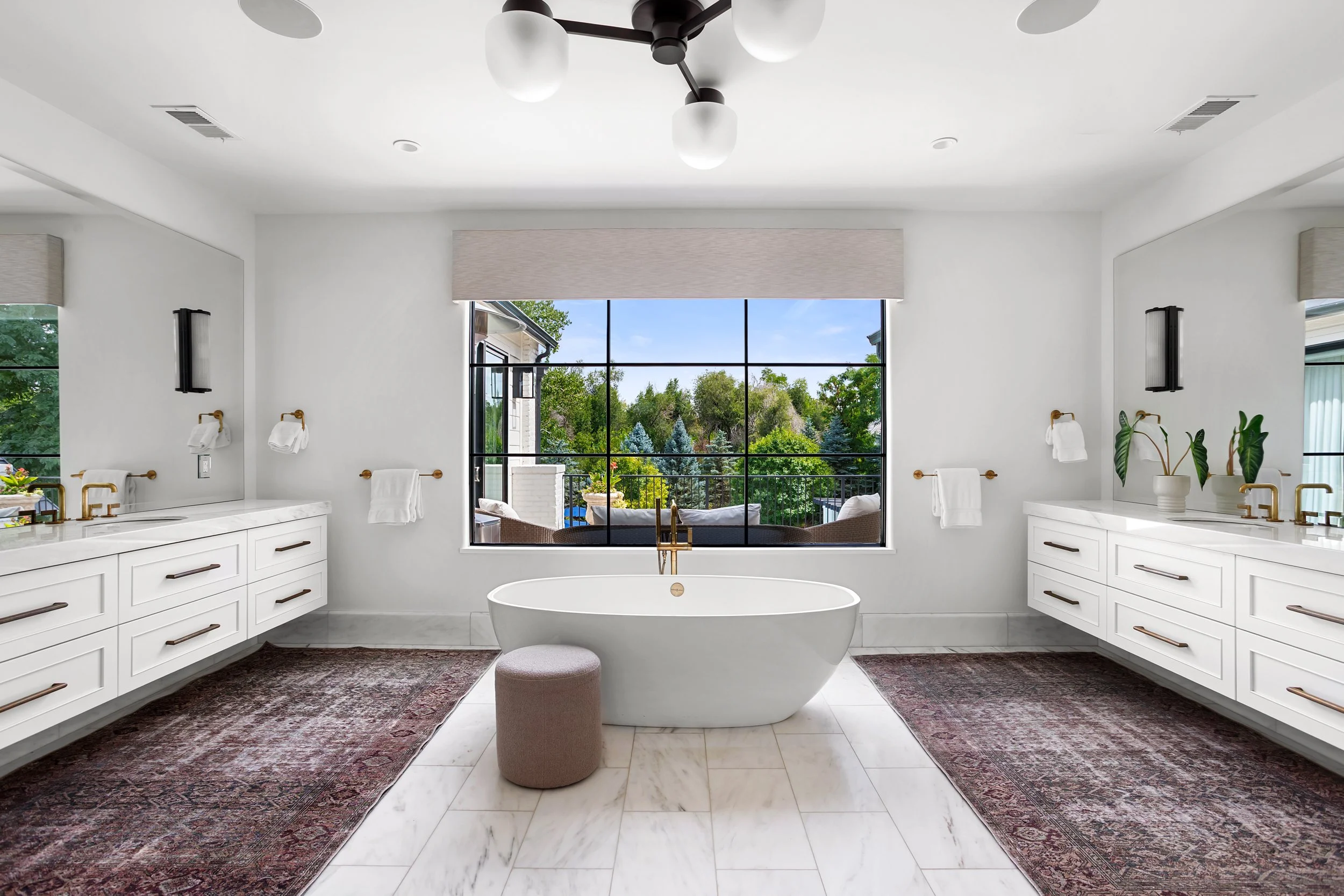 Bright bathroom with white cabinets, a freestanding bathtub, large window showing green trees outside, Persian-style rugs, and black wall-mounted lights.