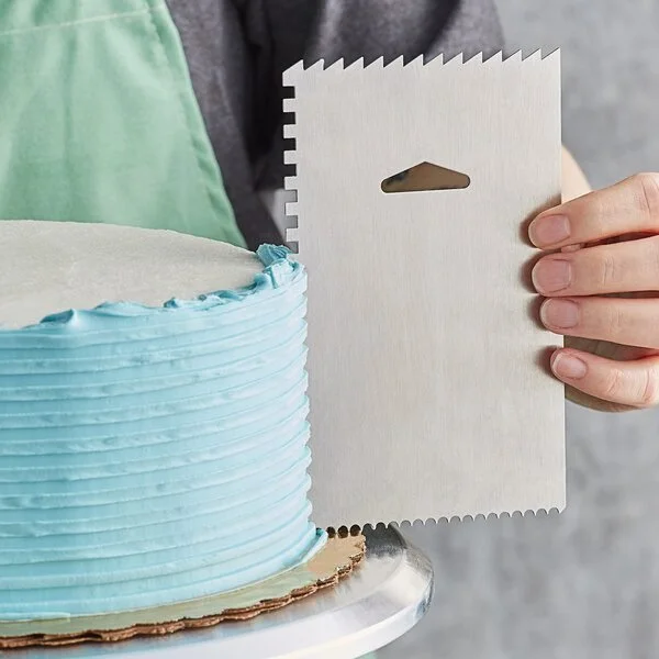Persona sostiene una espátula para cortar pastel con glaseado azul, al lado de un pastel decorado en tonos claros sobre un soporte plateado.