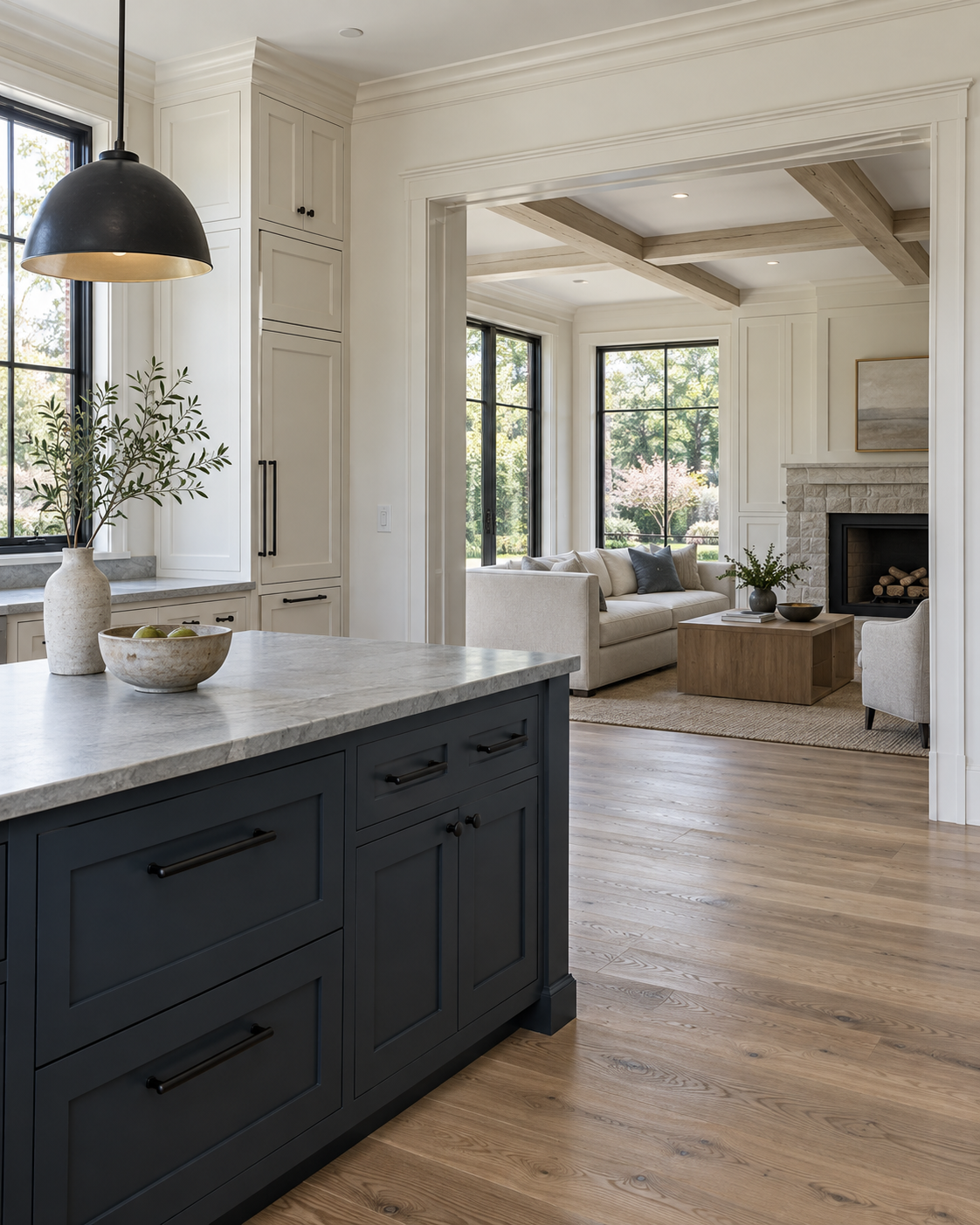 Open-concept living room with large windows, beige sofa, stone fireplace, wooden coffee table, and light hardwood flooring, viewed from the kitchen with navy blue cabinets and a marble countertop.