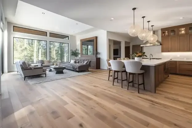 Open-concept living room and kitchen with large windows, modern gray sofas, wooden floors, and a kitchen island with barstools and pendant lighting.