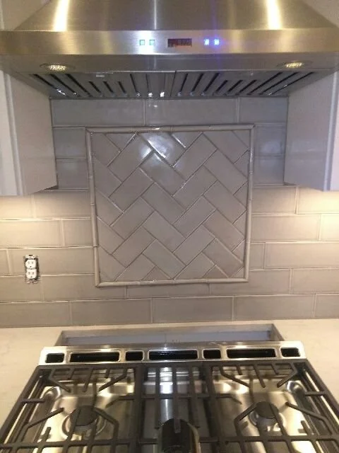 Kitchen stove with metallic range hood and tiled wall behind it.