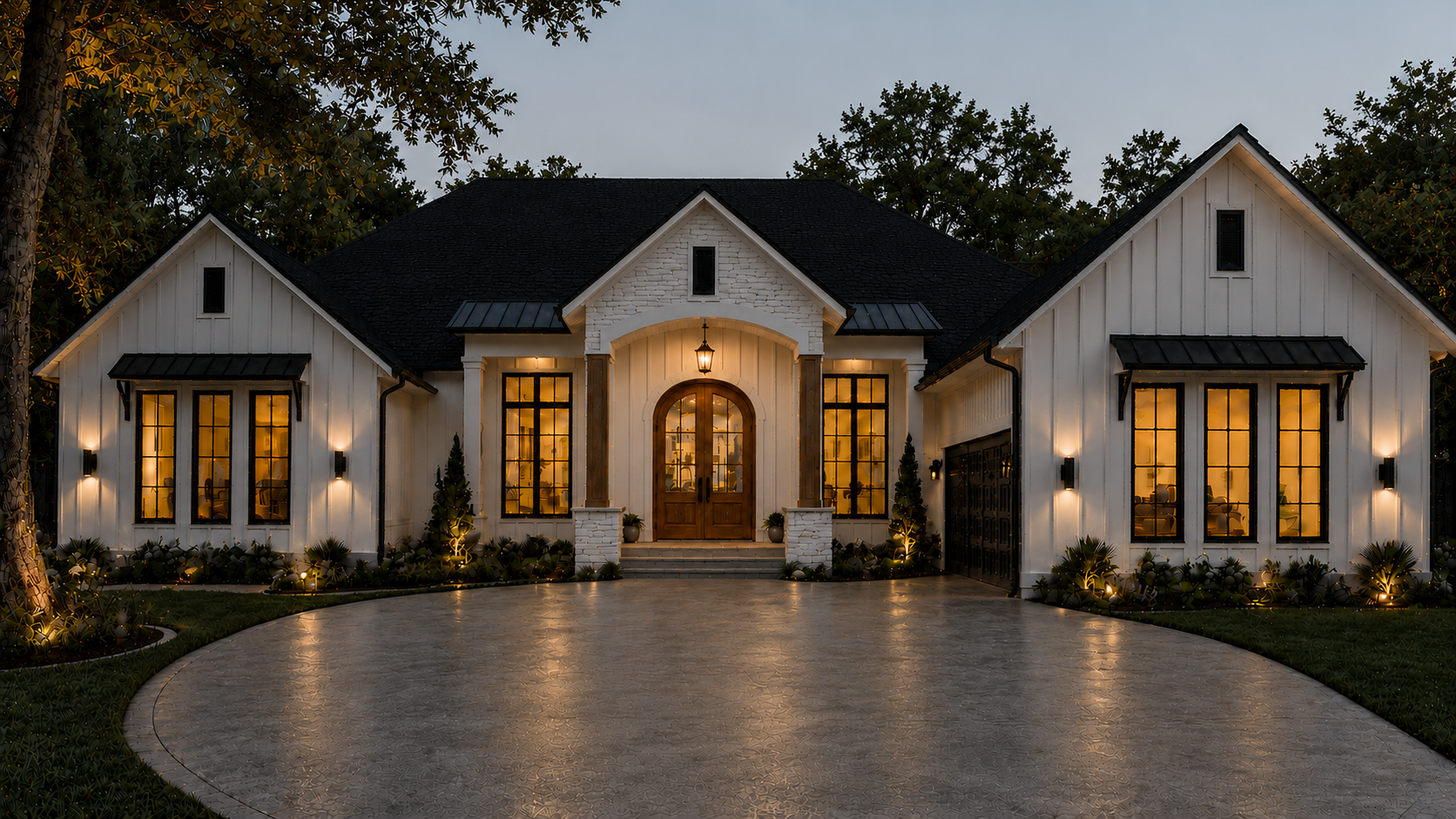 A modern white house with large windows illuminated from the inside during dusk or early evening. The house has a black shingle roof, with a central wooden door and steps leading up to it. There are exterior wall lights illuminating the front, and small decorative trees and plants with lights along the walkway. A curved driveway in front of the house reflects the warm interior lighting.