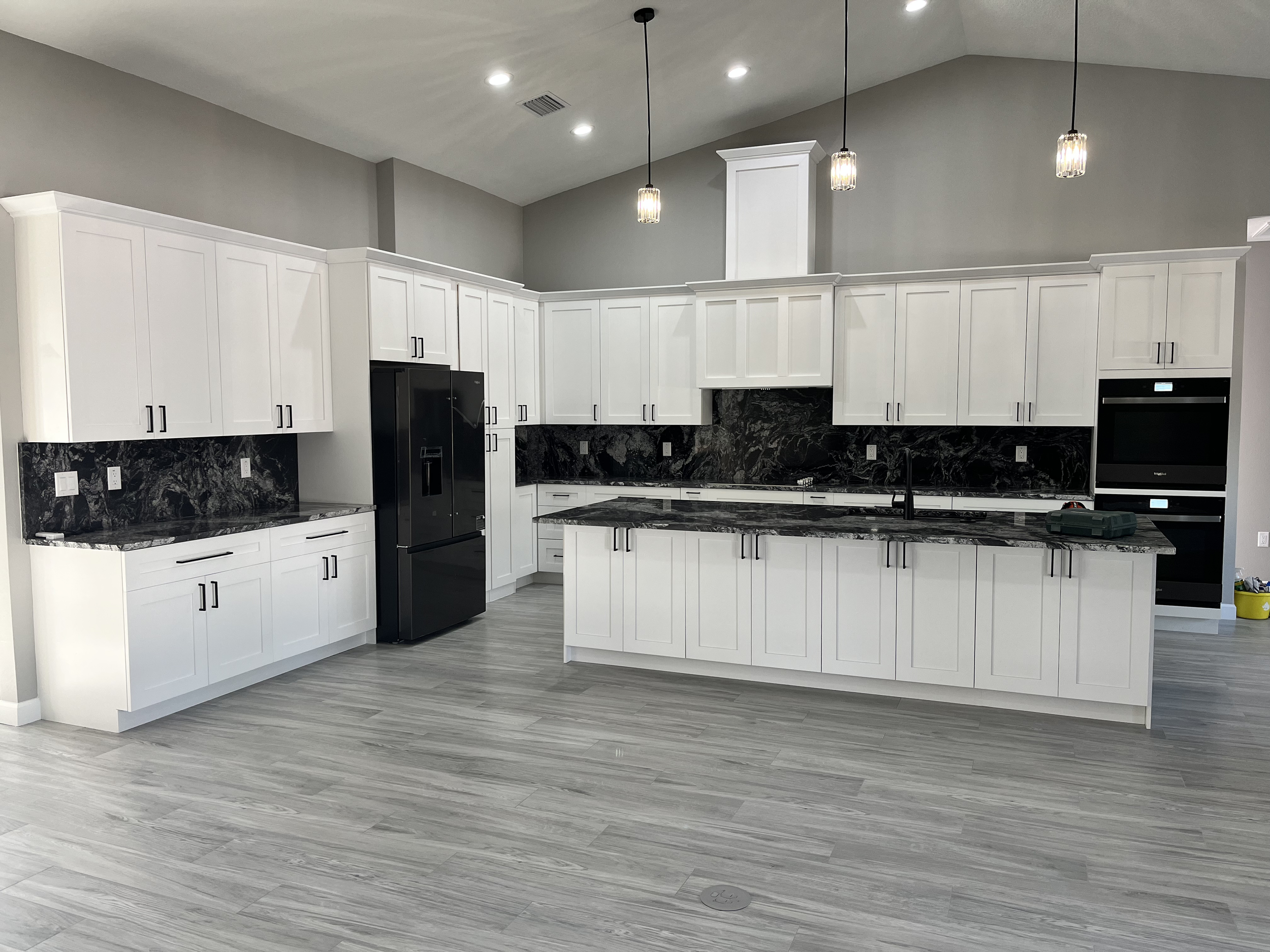 Modern kitchen with white cabinets, black appliances, and black granite countertops with white veining. Hanging pendant lights with glass shades are above the counter. The kitchen has a light gray wood floor, and a gray wall with a high ceiling.