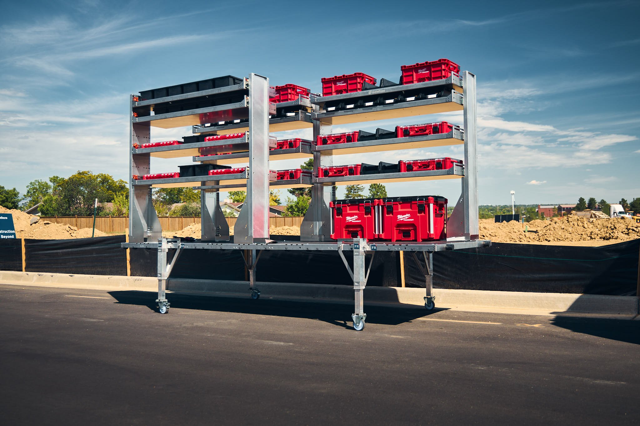 A large removable cargo van upfitting system with shelves and red tool boxes, set up on an asphalt surface at a construction site with dirt and fencing in the background, under a blue sky with scattered clouds.