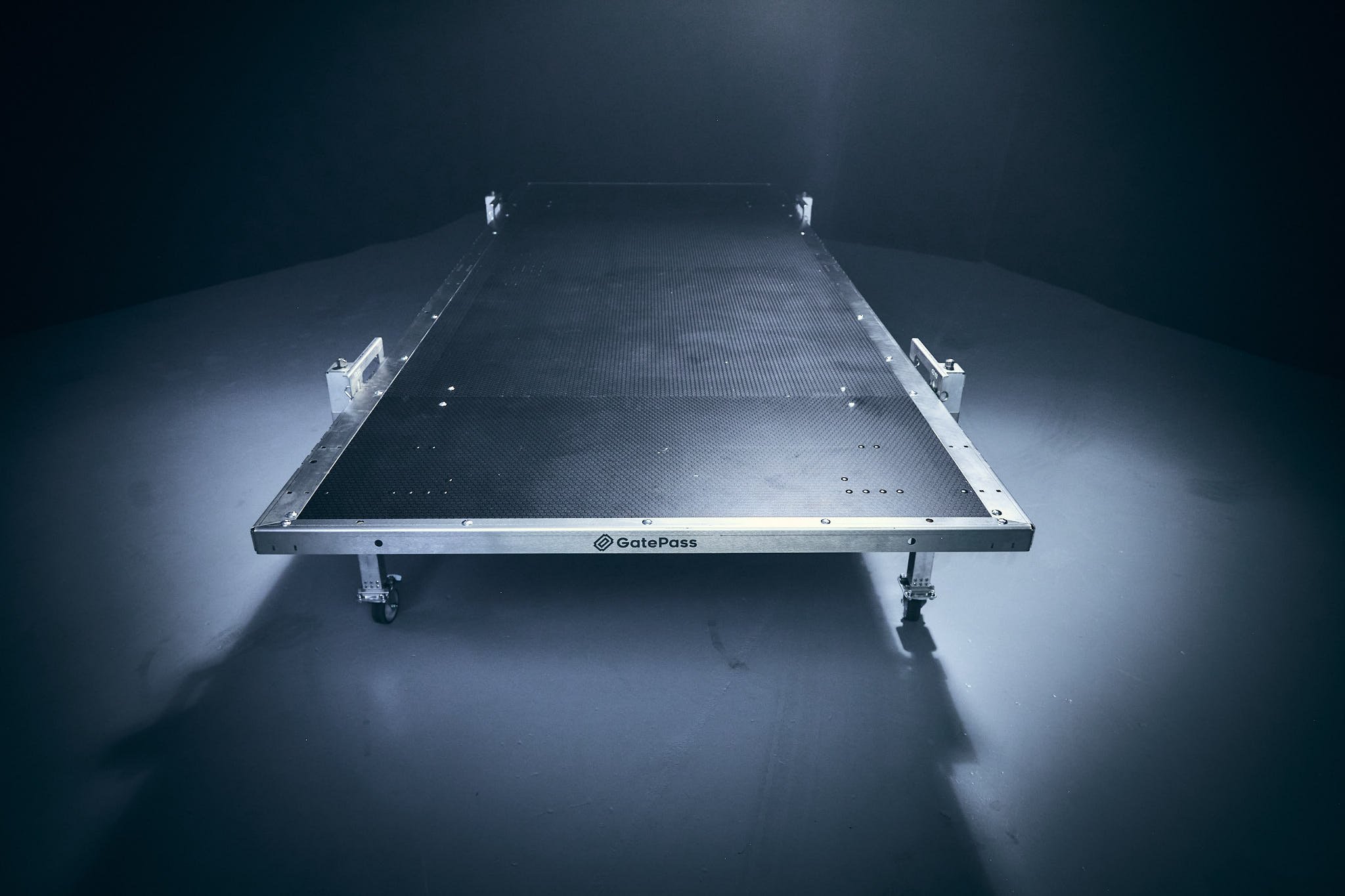 A black, metal platform on wheels with a textured surface and small white markings, labeled 'GatePass,' positioned in a dark room with lighting focused on it.