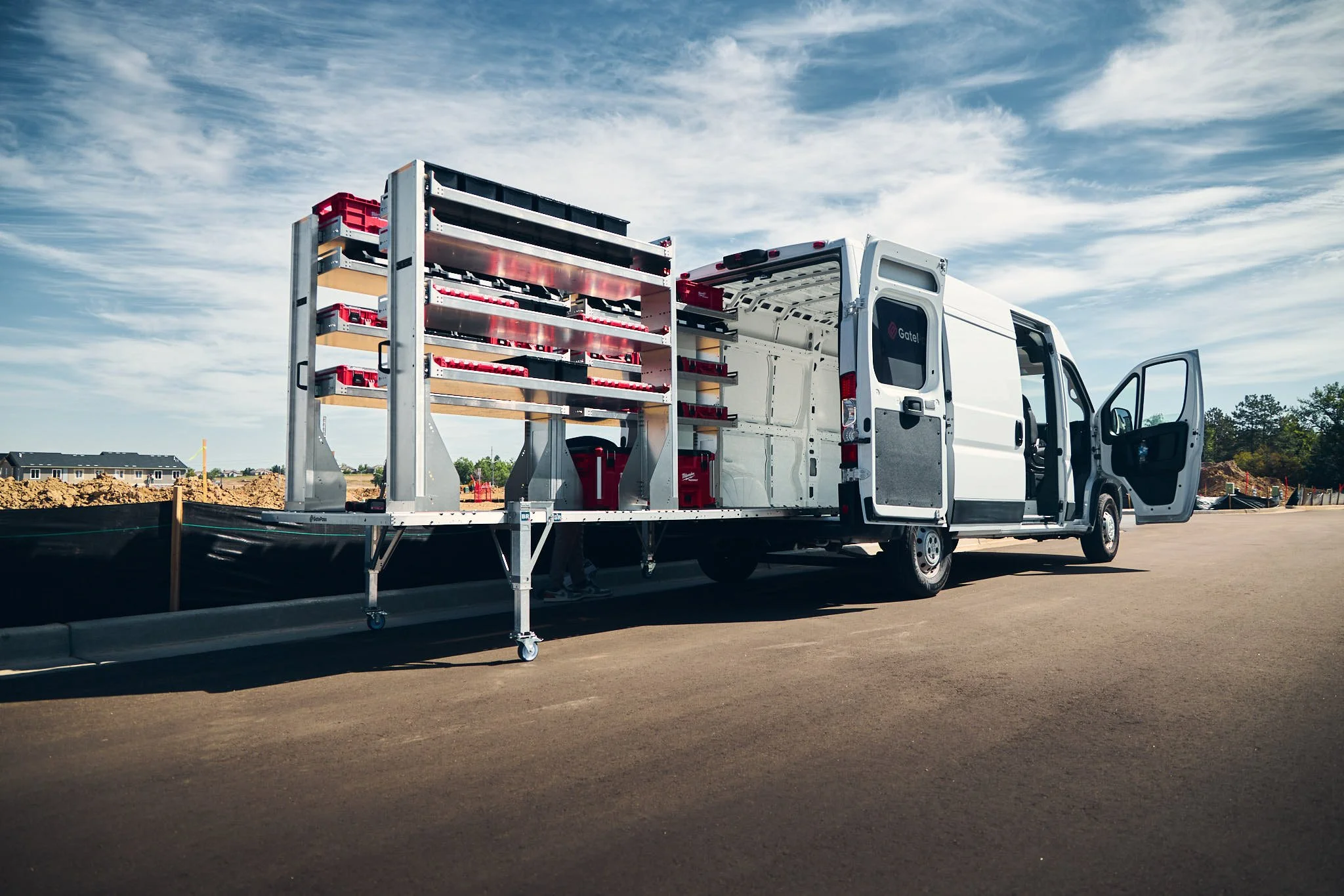A white work van with its side door open parked on a construction site. The van has a large removable cargo van upfitting system with shelves and red tool boxes behind it. The sky is partly cloudy.