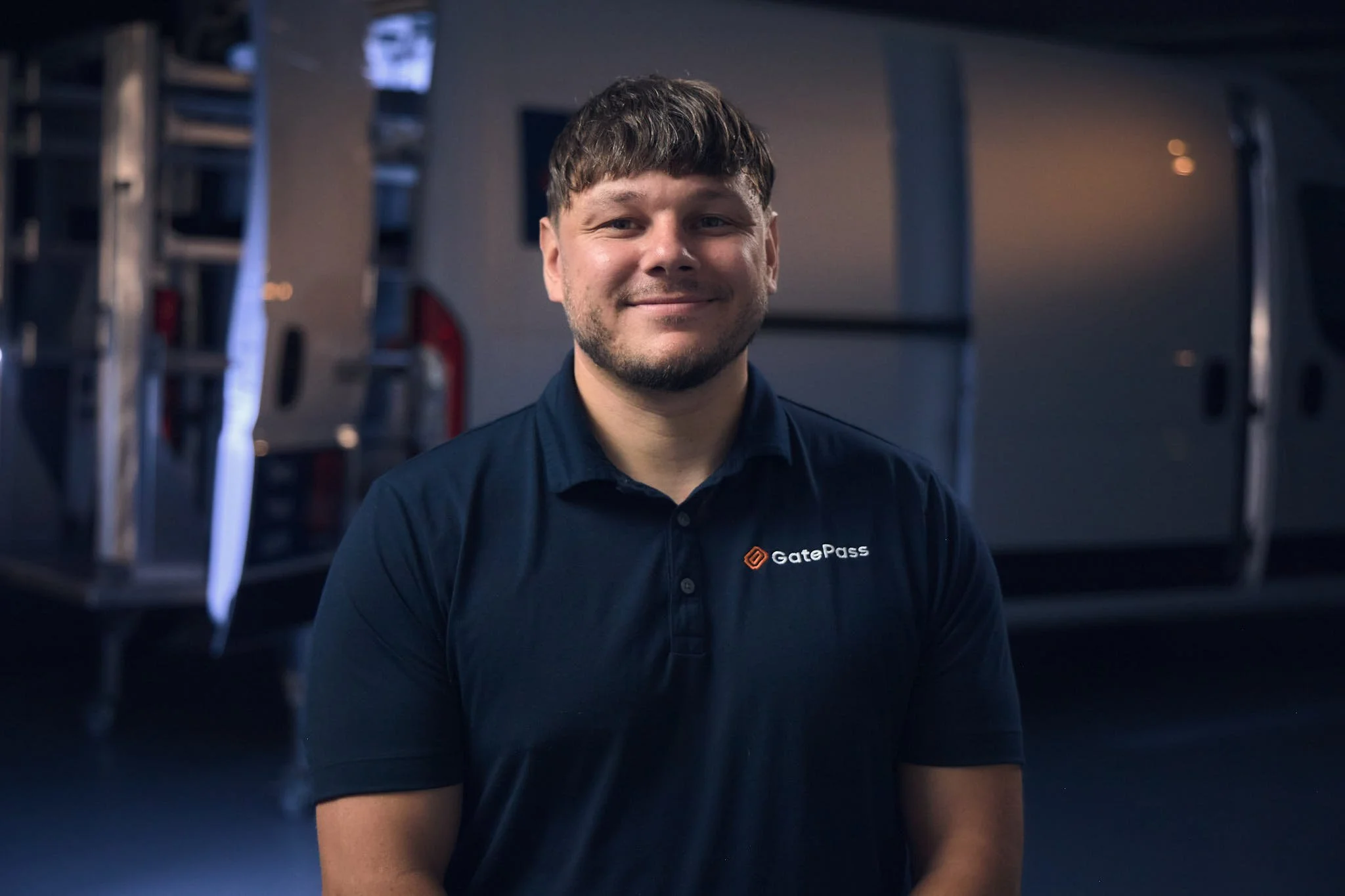A young man with short brown hair and a beard smiling in front of a blurred industrial background, wearing a black shirt with the logo 'GatePass'.