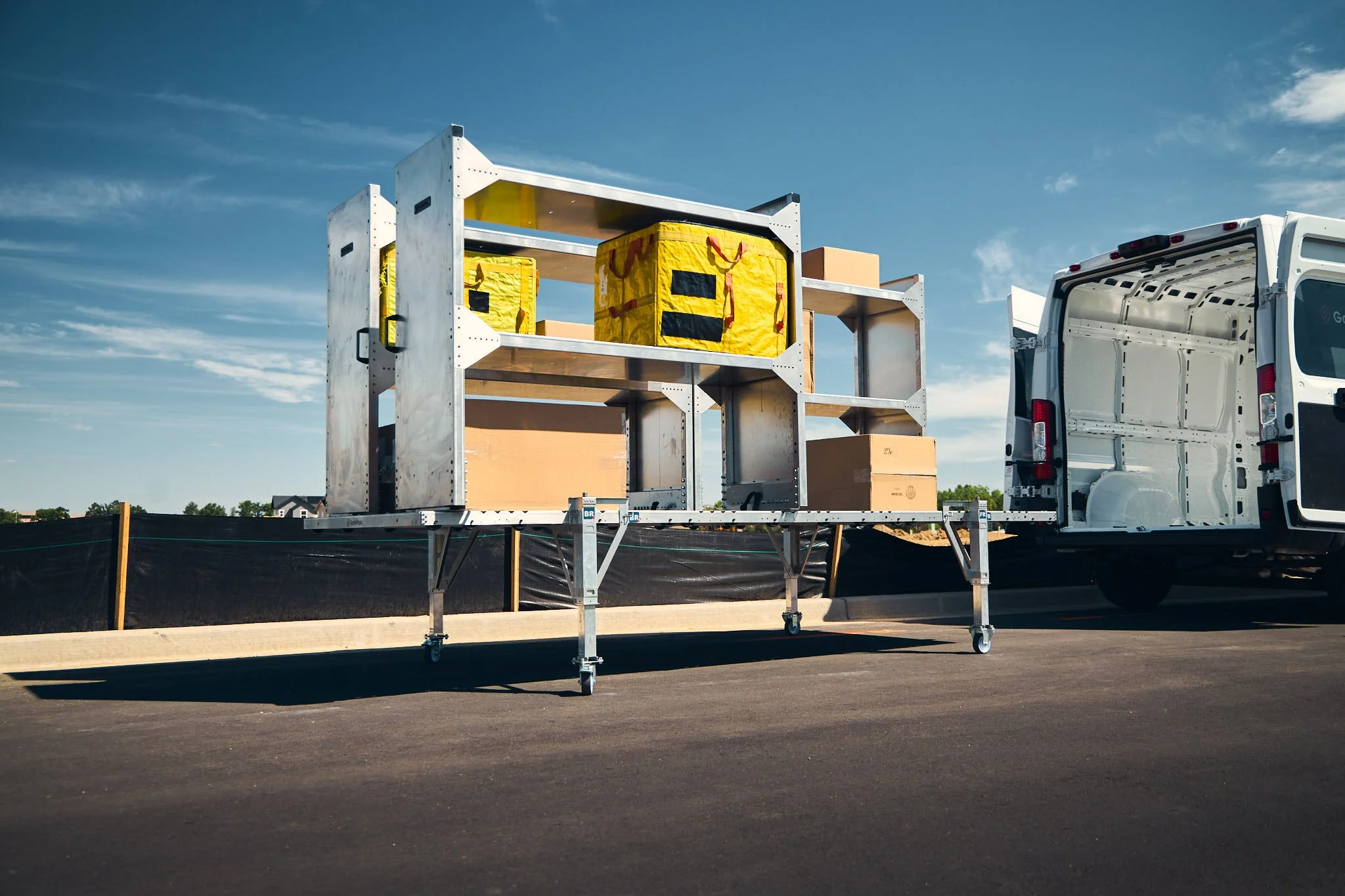 A large removable cargo van upfitting system with shelves loaded with cardboard boxes and yellow insulated delivery bags, parked on a paved surface under a clear blue sky.