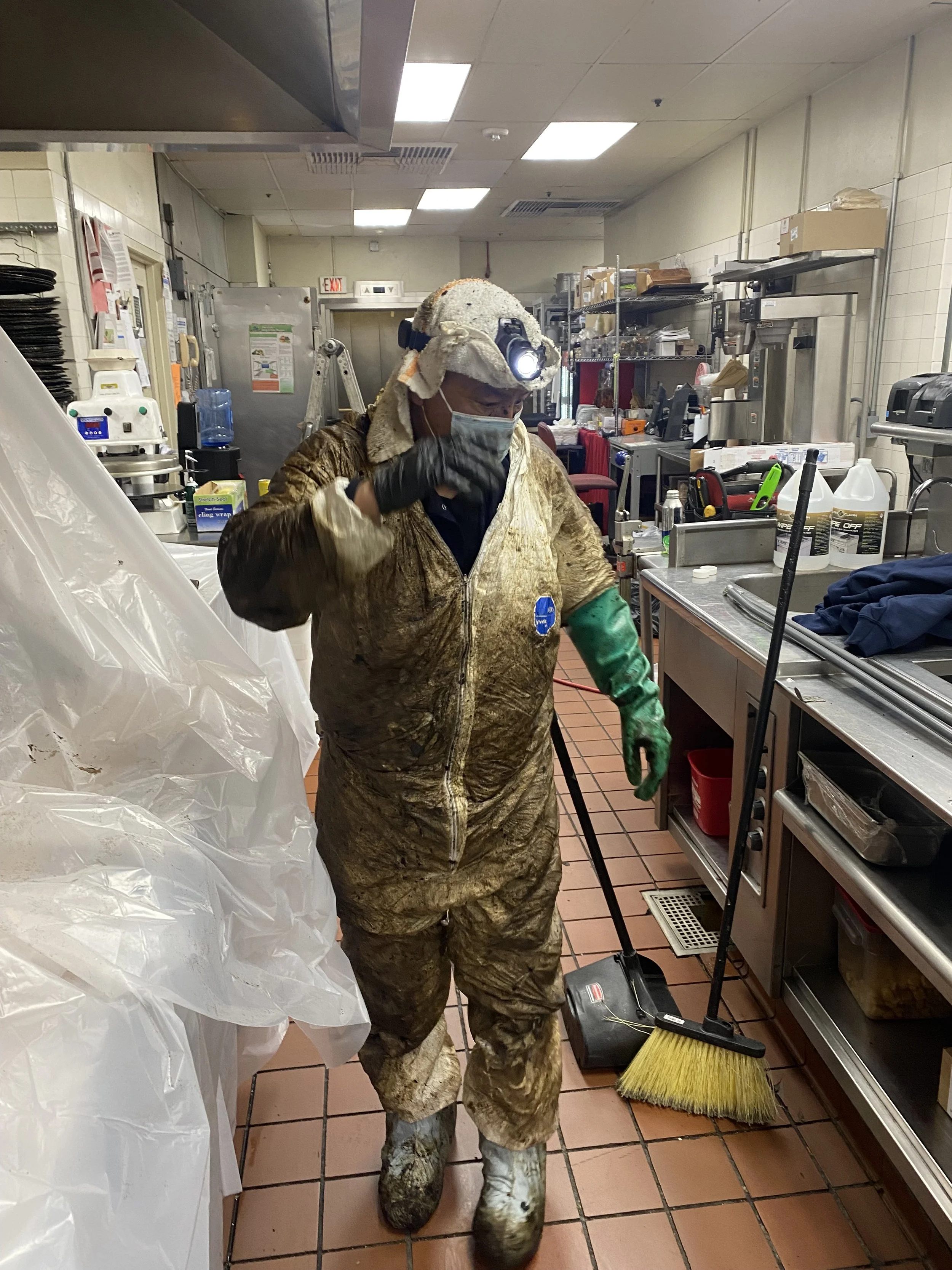A worker in a commercial kitchen wearing a protective suit, face mask, gloves, and a headlamp, using a broom to clean the tiled floor.