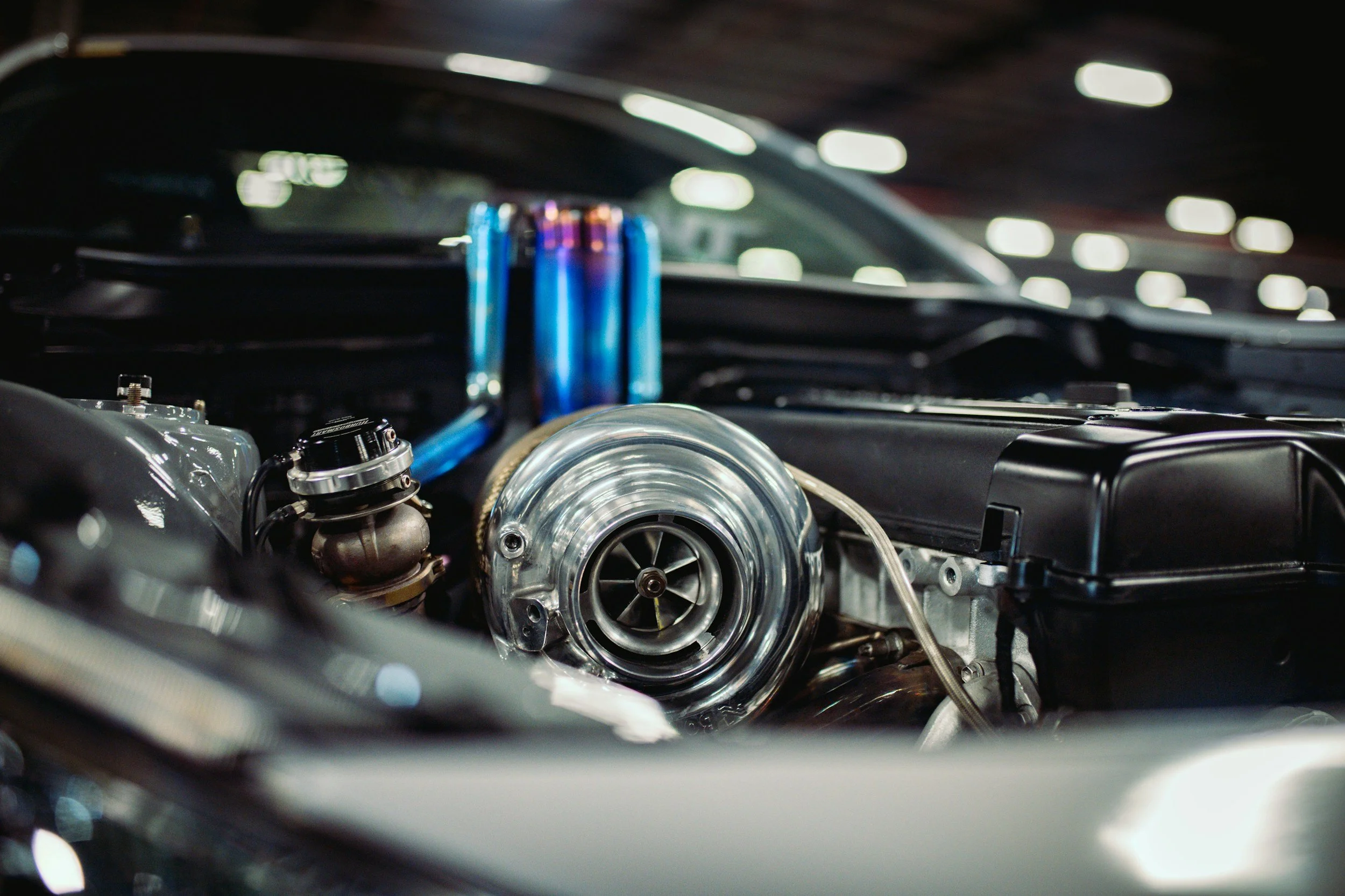 Close-up of a car engine with turbocharger and blue and purple parts inside the engine bay, under bright indoor lighting.