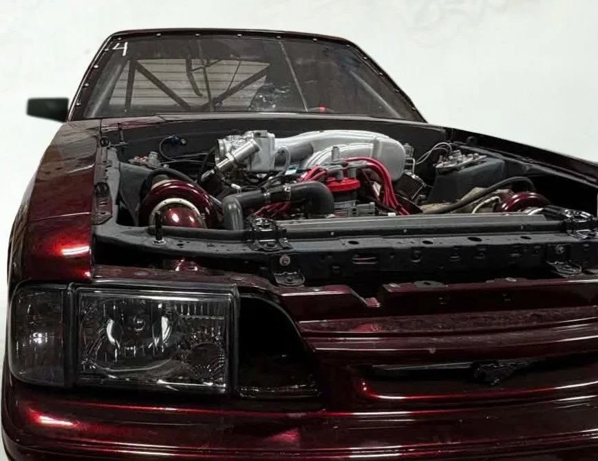 Close-up of a vintage car with its hood open, revealing a high-performance engine with red and silver components.