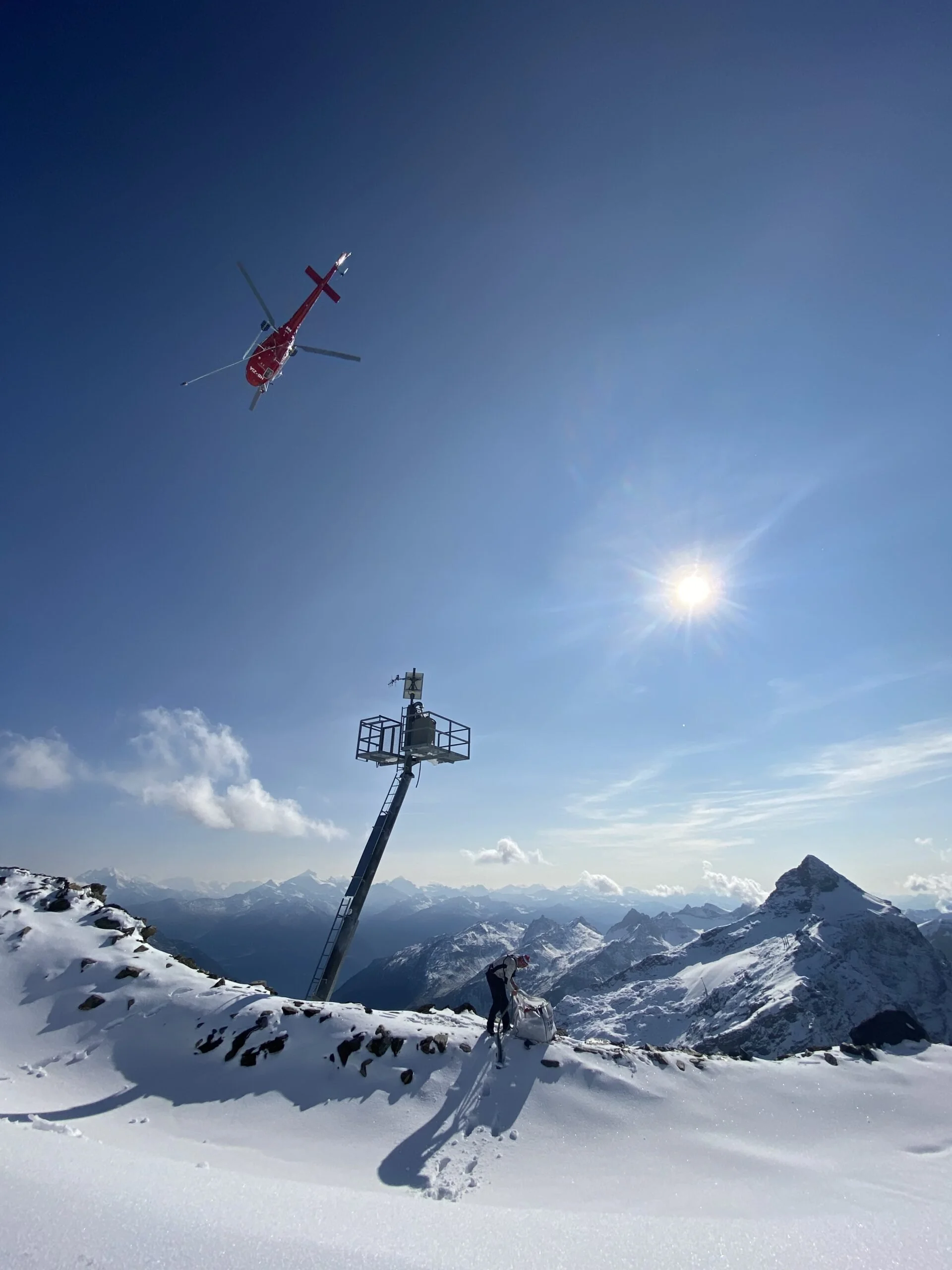 Snow-covered mountain landscape with a person using a sled, a communication tower, and a helicopter flying in the blue sky with the sun shining behind clouds.