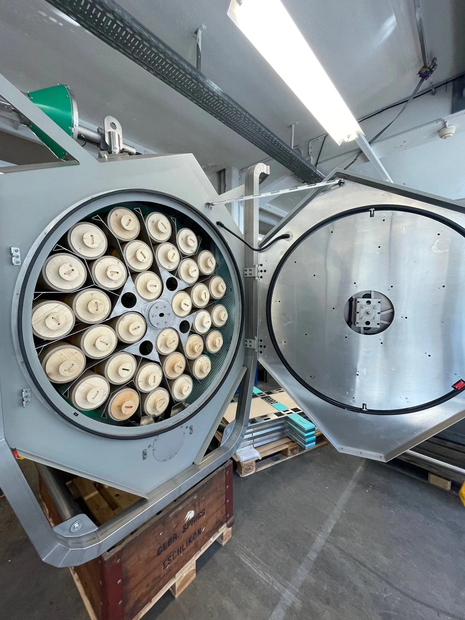 Industrial machine with a large circular component containing multiple smaller beige rollers, attached to metallic framing and situated on a wooden pallet in a warehouse.
