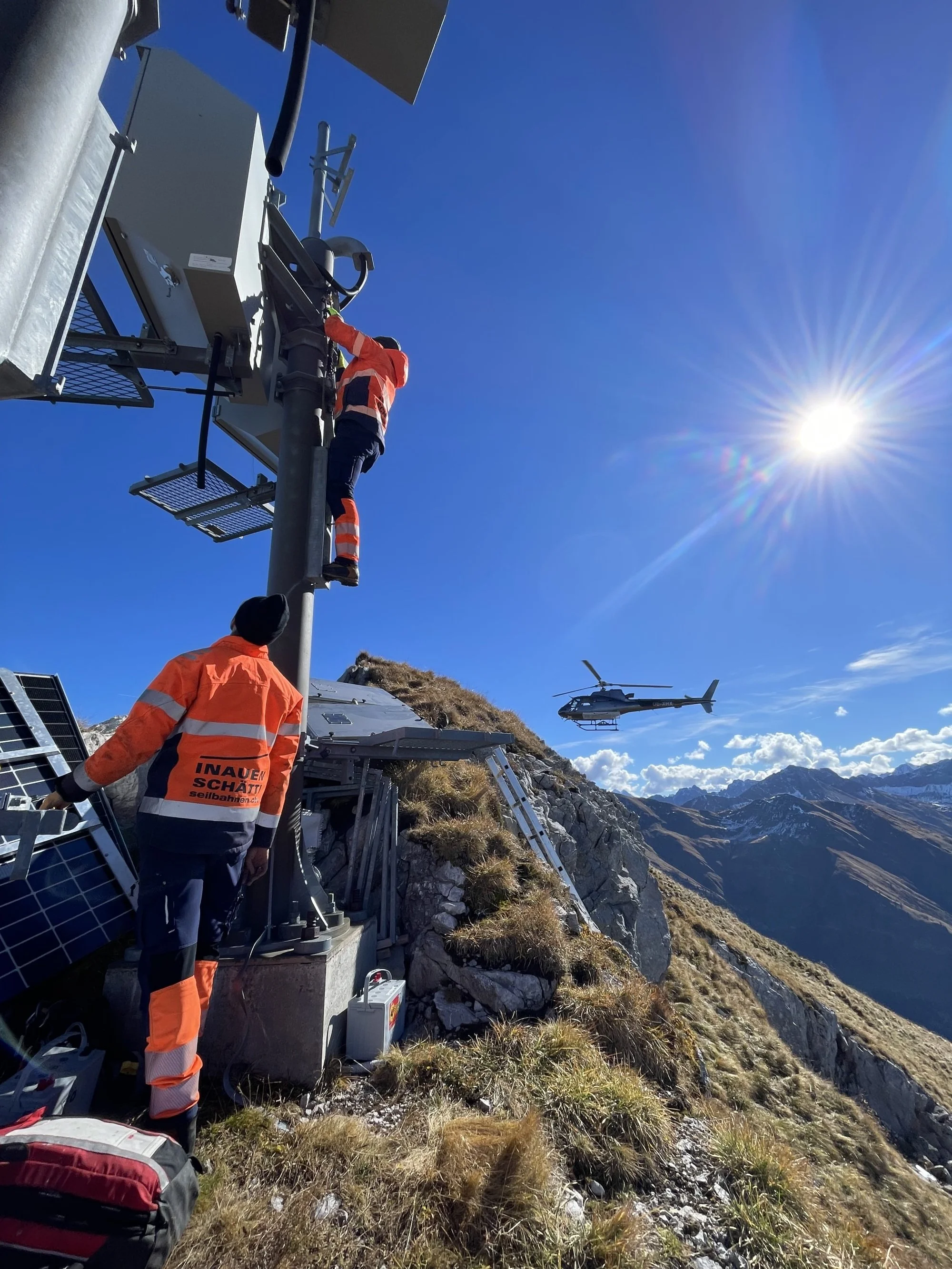 Three workers in orange safety clothing are working on equipment on a mountain slope. A helicopter is flying in the clear blue sky, and there's a scenic mountain landscape in the background.