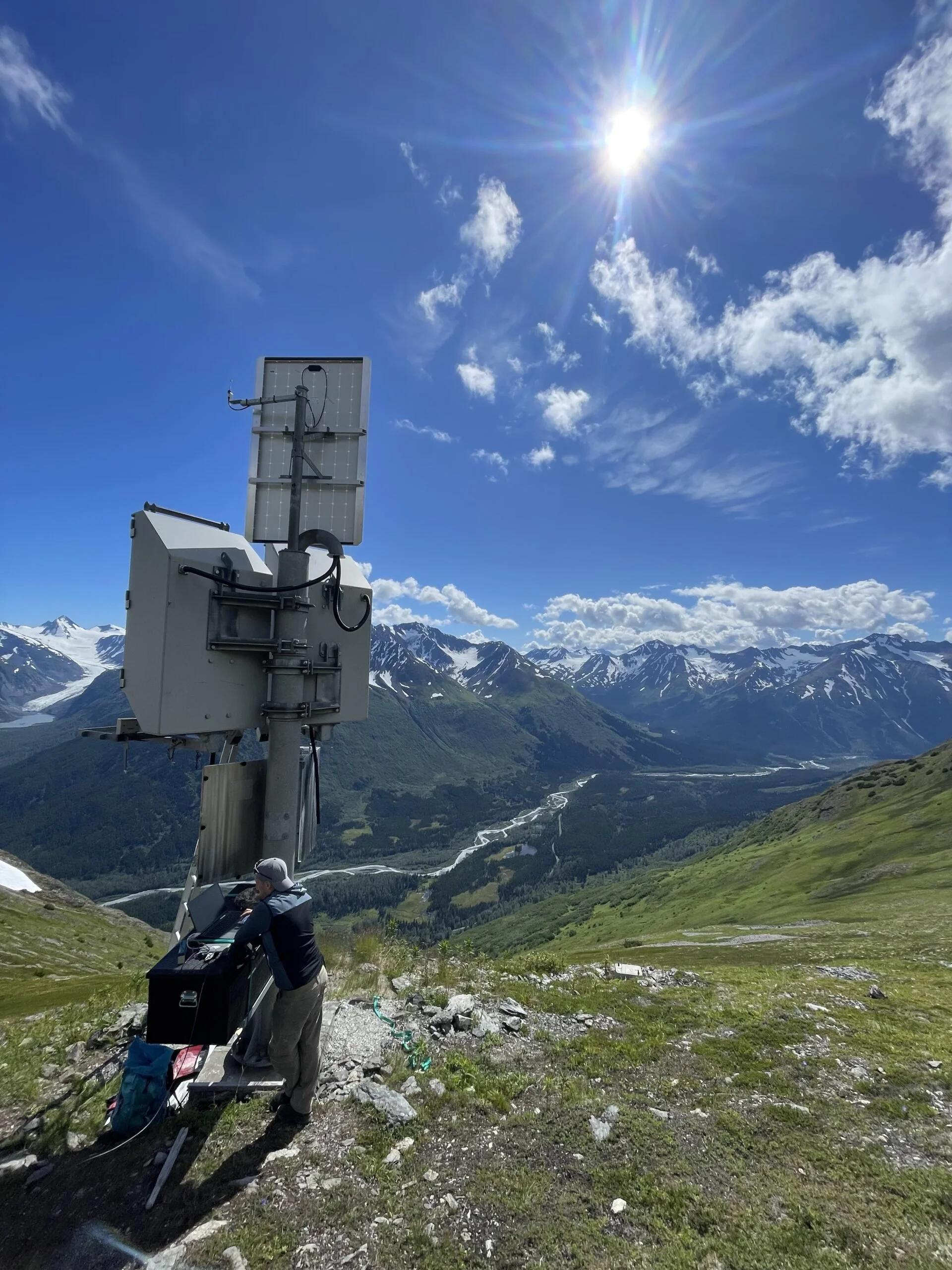 Scientists setting up a solar panel telescope in a mountainous landscape under a bright sunny sky with some clouds.
