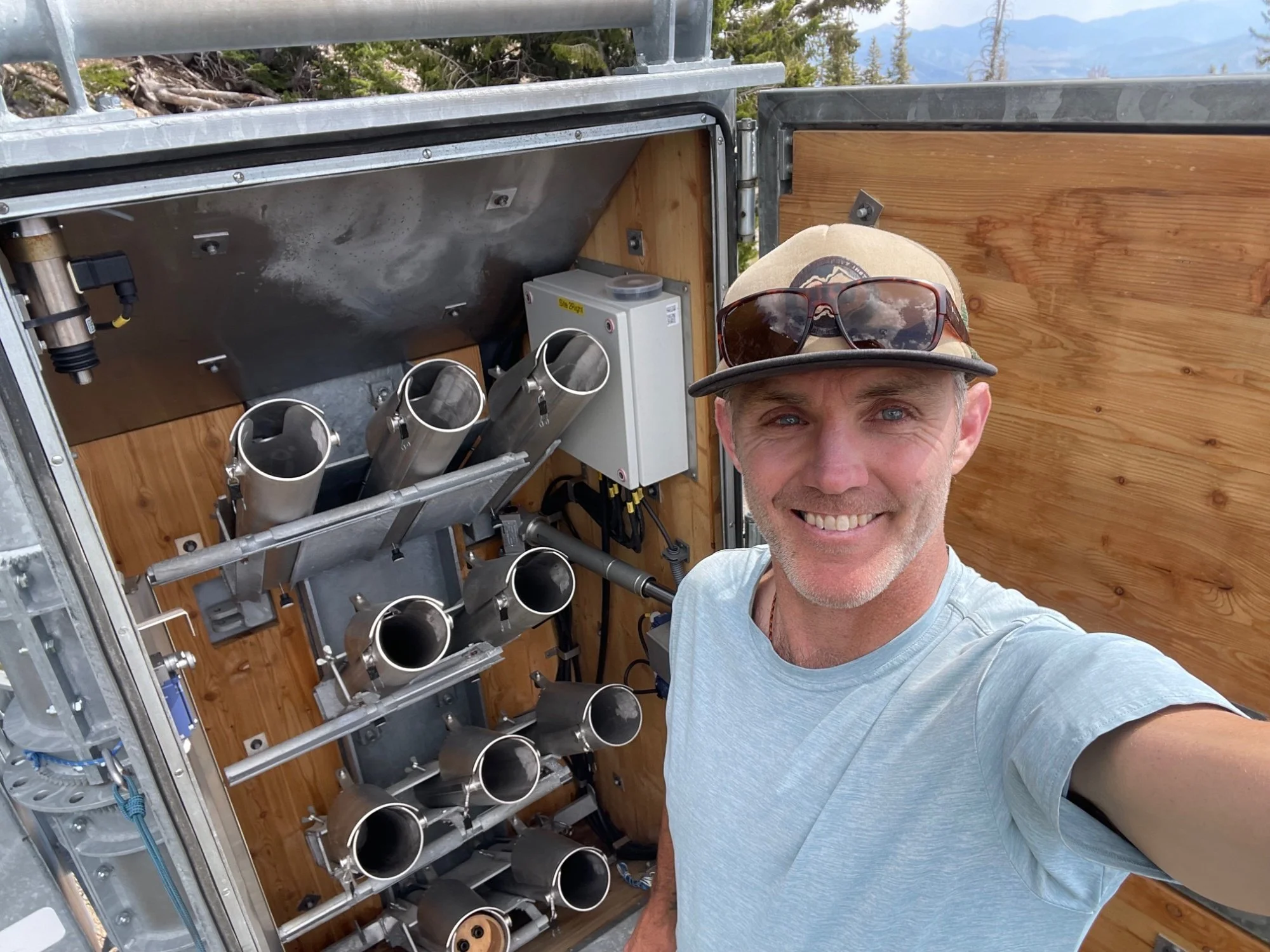 A man smiling in a light gray t-shirt and beige cap with sunglasses on top takes a selfie in front of a metal and wood structure that holds multiple mechanical or electronic components, with a mountain landscape in the background.