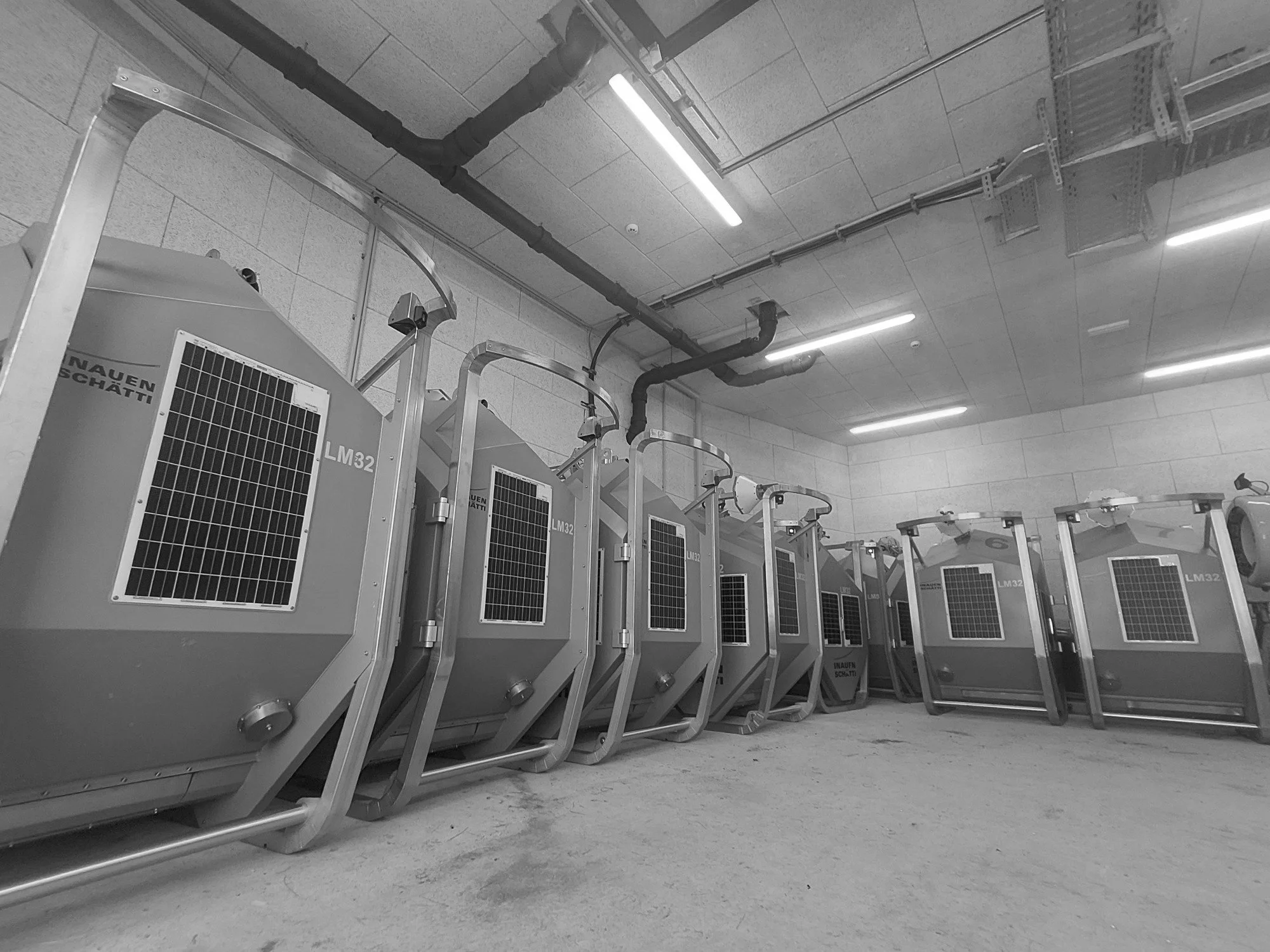 Row of industrial air conditioning units in a basement with exposed pipes and fluorescent lighting.