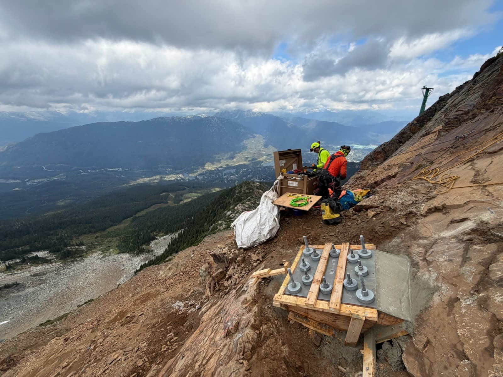 Two workers in safety gear performing geological or engineering work on a mountain slope with a scenic valley and mountains in the background.
