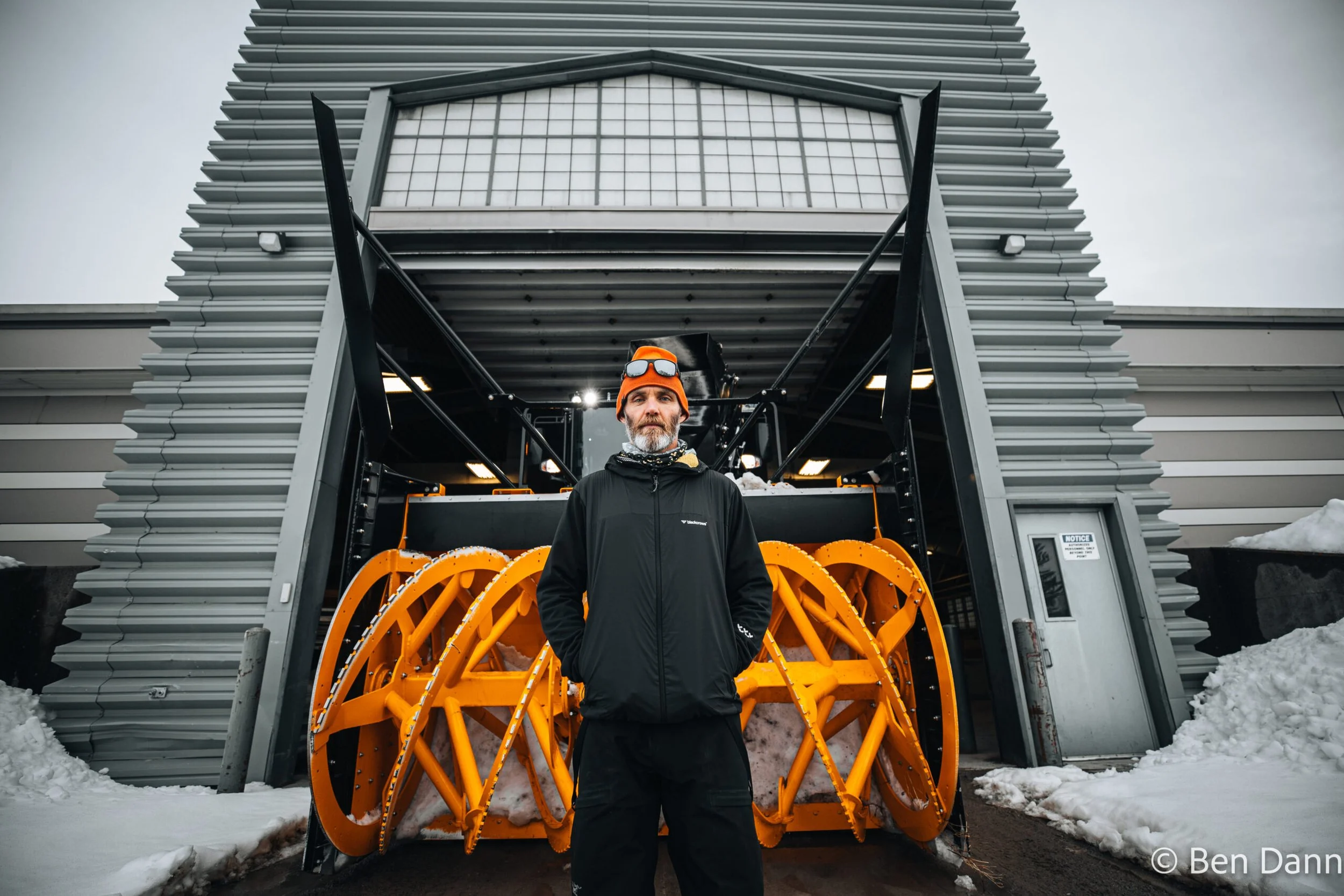A man standing in front of a large snow grooming machine outside a modern building with horizontal siding. The man is wearing a black jacket, an orange beanie, and sunglasses on his forehead. Snow is on the ground and piled on the sides.