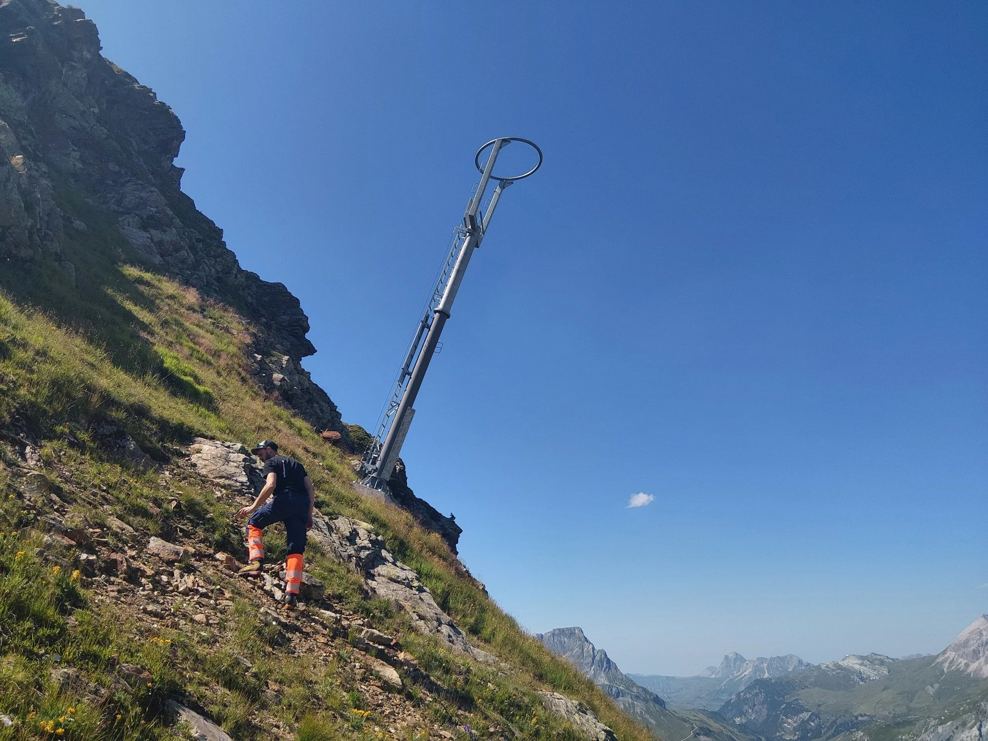 A hiker walking up a steep rocky hillside with a mountain landscape in the background, a blue sky, and a tall metal pole