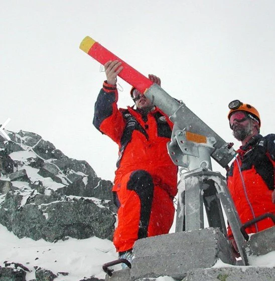 Two climbers in orange gear using a mountaintop telescope in snowy terrain.