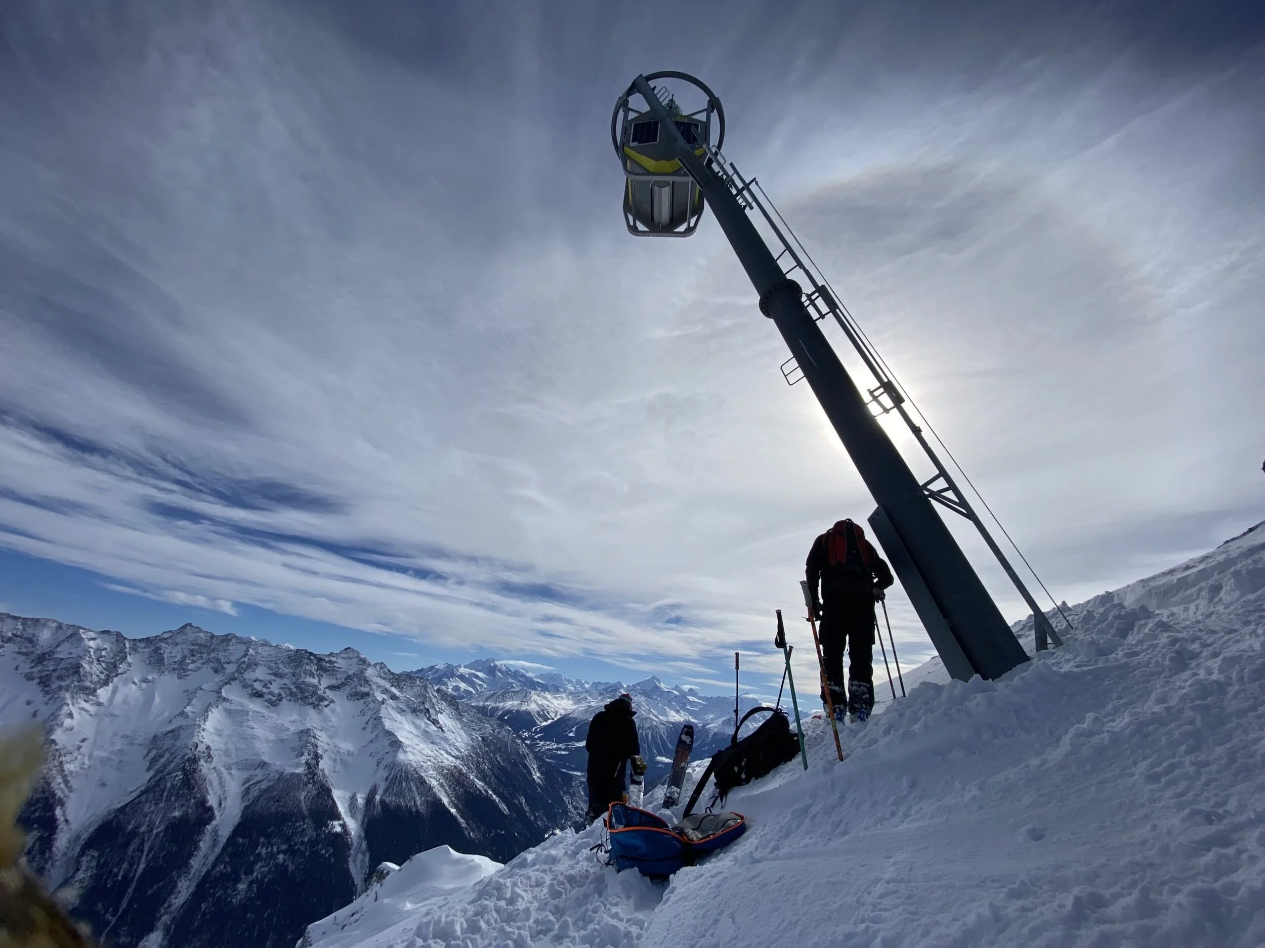 Snowy mountain landscape with a snow-covered slope, mountain peaks in the distance, and clear sky with some clouds. Several people are visible, some wearing ski gear, and a tall structure with a basket like attachment at the top is present.
