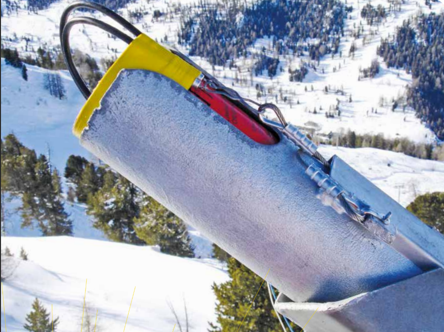 Close-up of a snow probe or tool with a yellow handle, metal shaft with snow and ice, set against a snowy mountain landscape with trees in the background.