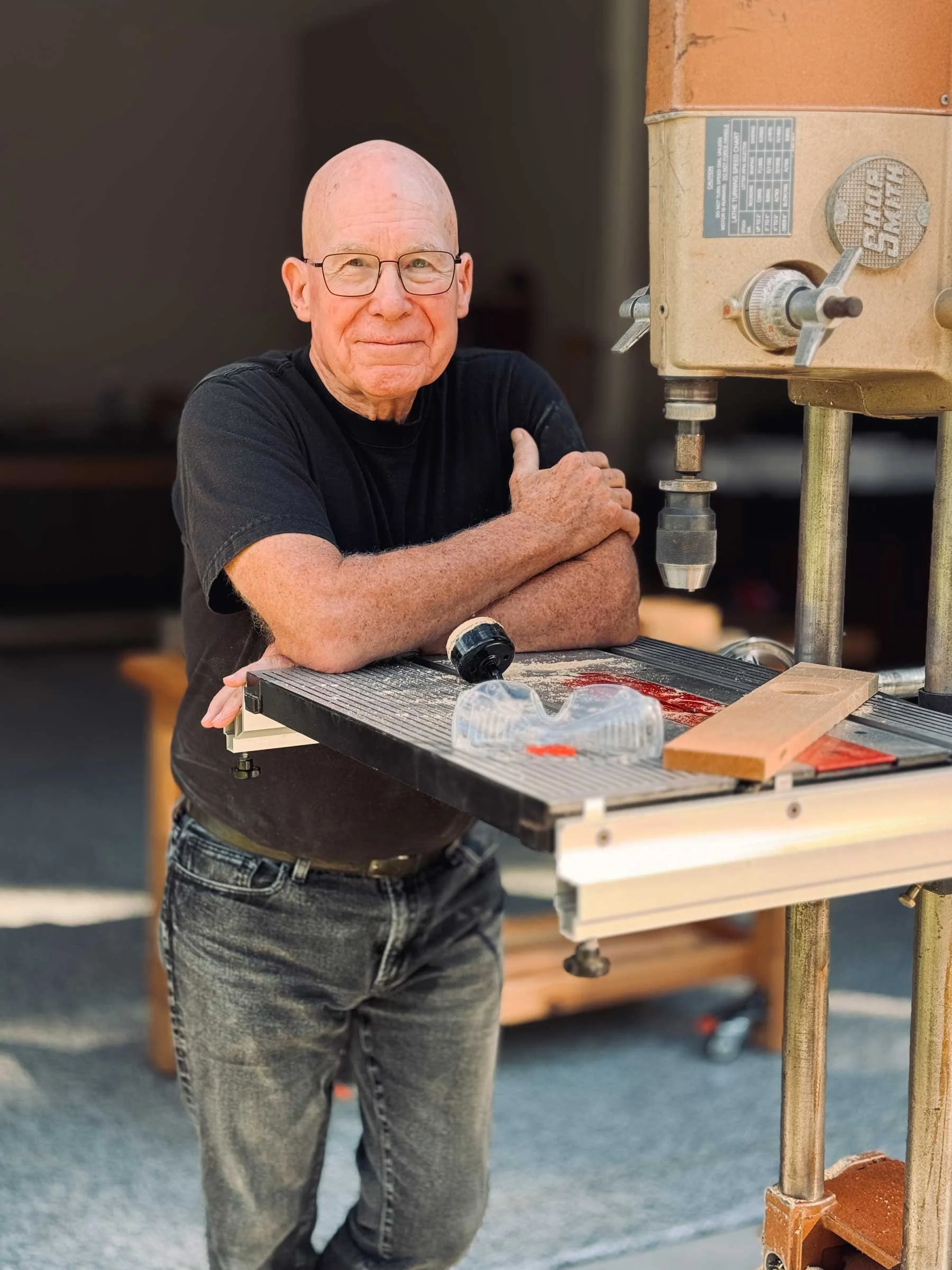 An elderly man with glasses, wearing a black T-shirt and gray jeans, leaning on a table with a woodworking drill press nearby. The background shows a blurred outdoor setting.