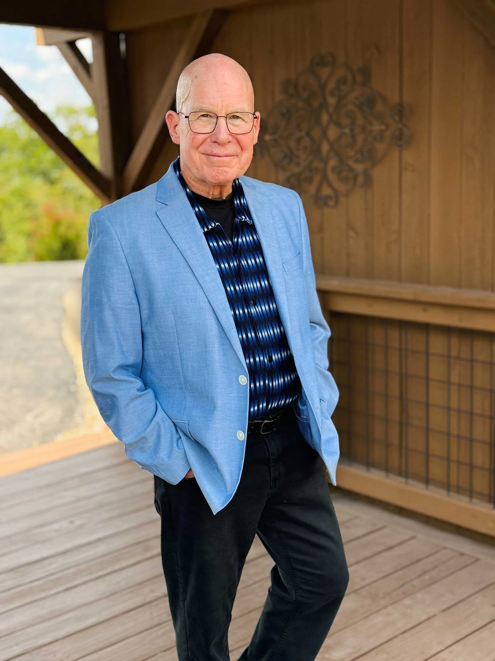 Portrait of a smiling bald man wearing glasses, a blue blazer, a patterned blue shirt, and black pants, standing outdoors on a wooden deck with a background of trees and a wooden wall with decorative metalwork.