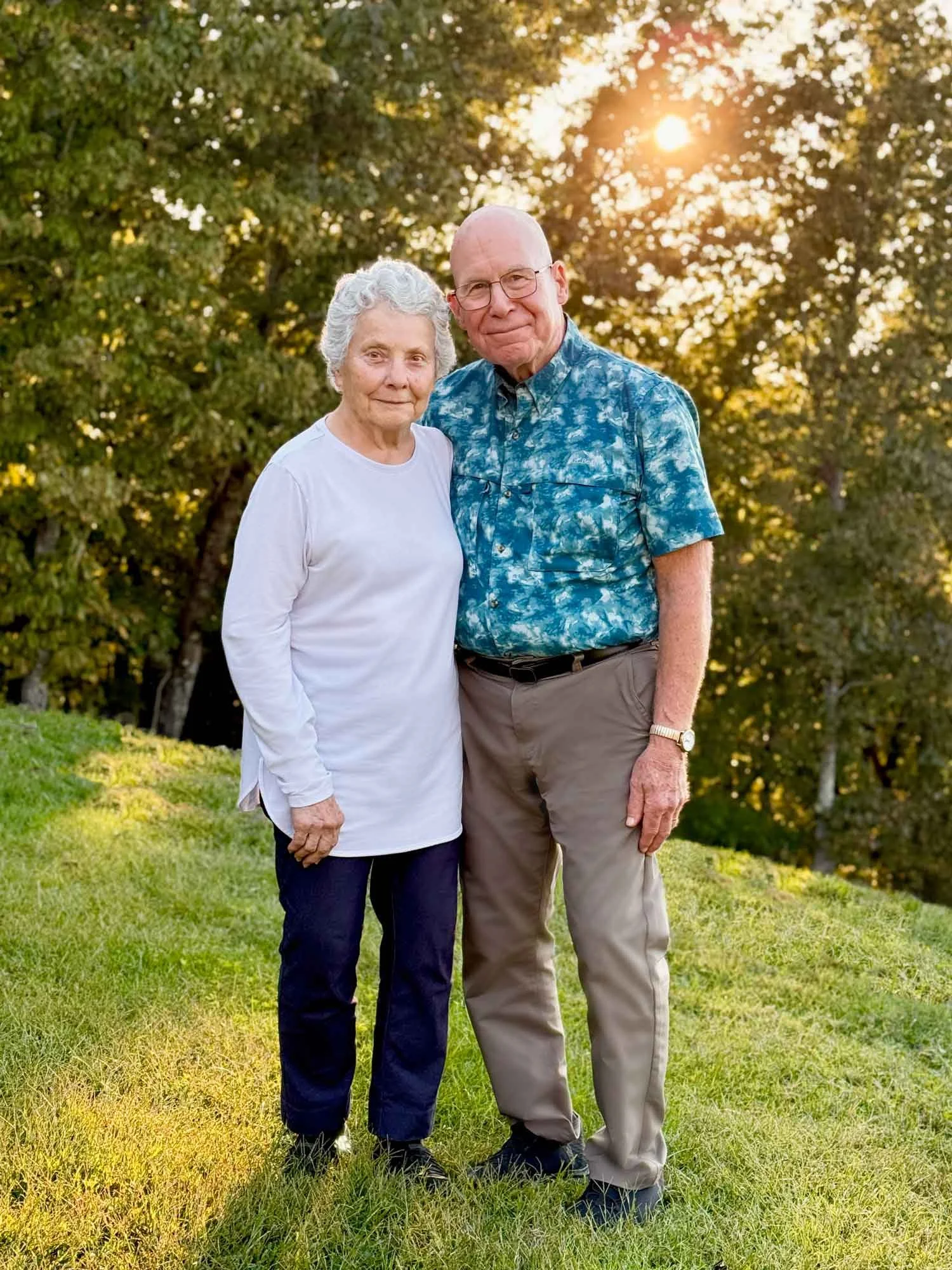 An elderly woman and man standing outdoors on green grass with trees in the background during sunset.