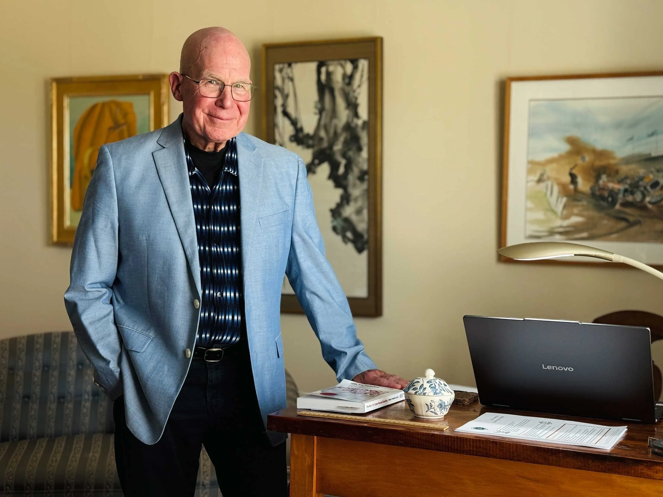 An elderly man wearing glasses and a blue blazer standing in a room with framed artwork on the wall. He is smiling and standing next to a wooden table with books, documents, a decorative bowl, and a laptop.