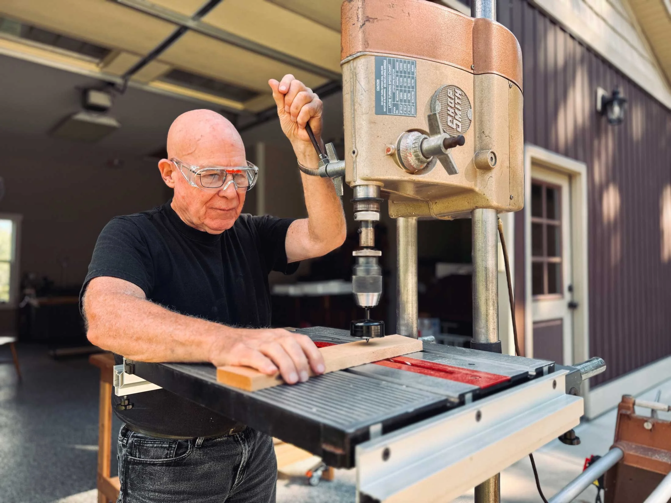 A man with glasses operating a woodworking drill press outdoors in the daytime.
