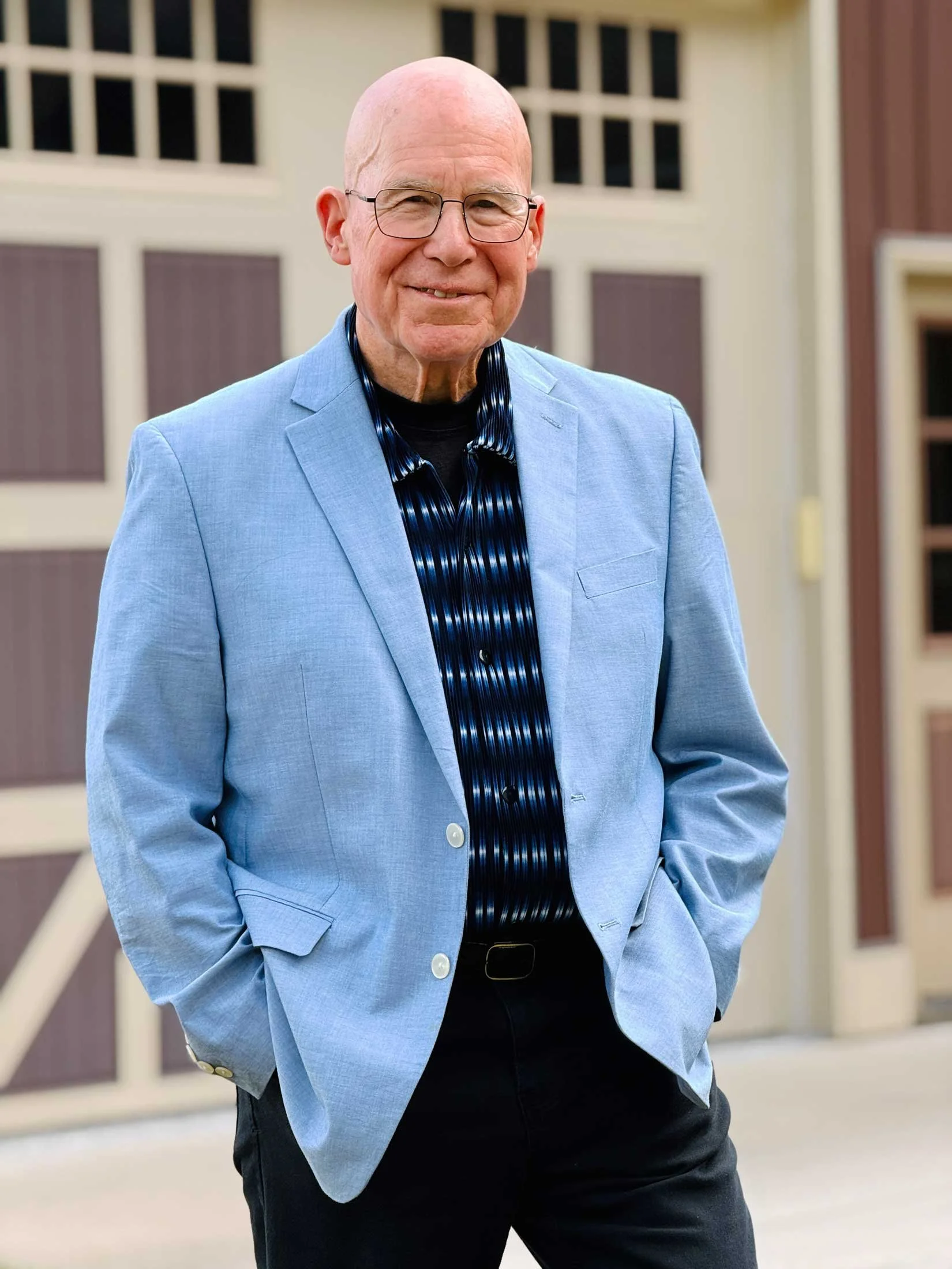 A smiling bald man with glasses wearing a light blue blazer over a black shirt with a blue pattern, standing outside in front of a building with windows.