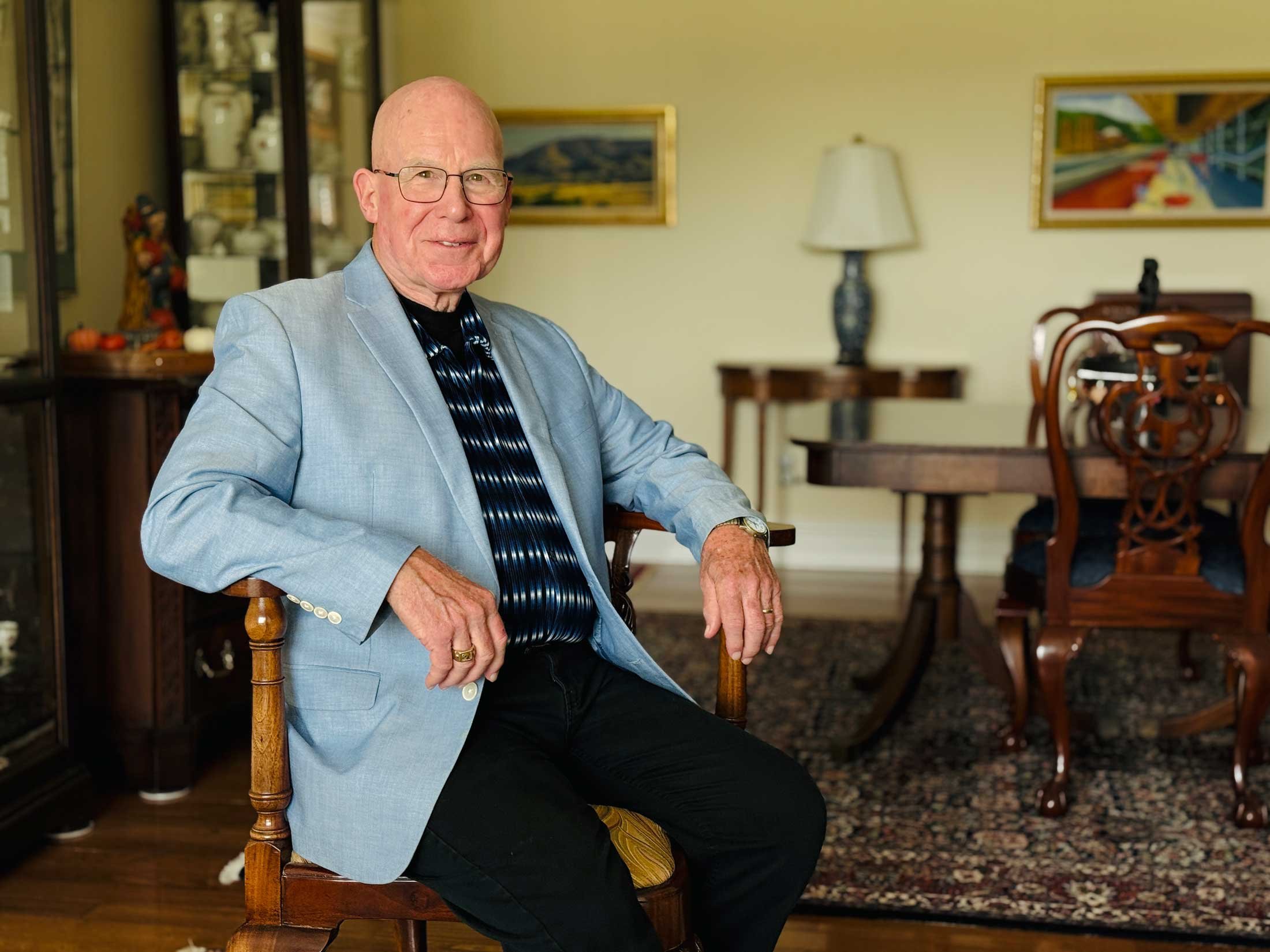 An elderly man with glasses and a light blue blazer sitting on a wooden chair in a dining room. He is smiling slightly with one hand resting on his lap. The background includes a table with chairs, artwork on the wall, and a lamp.
