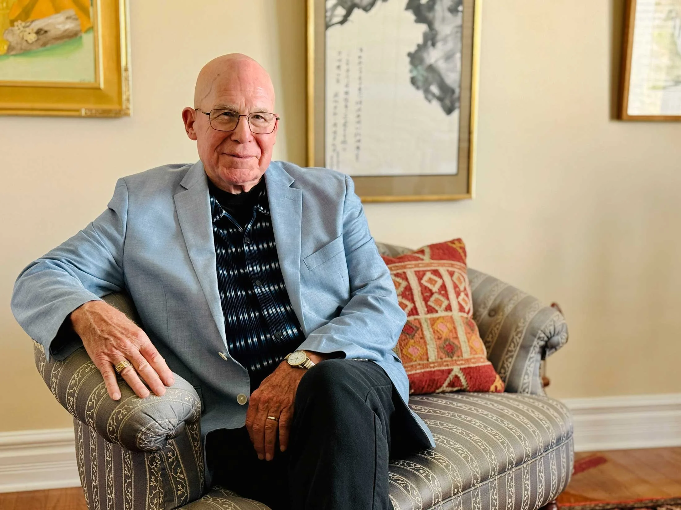 An elderly man with glasses, wearing a light gray blazer over a dark, patterned shirt, sits on a patterned beige armchair with ornate details. He has a watch on his left wrist and a ring on his left ring finger. Behind him, there is a decorative pillow with a red and beige geometric pattern on a matching sofa. There are framed paintings with landscapes and traditional Asian art on the beige wall behind him.