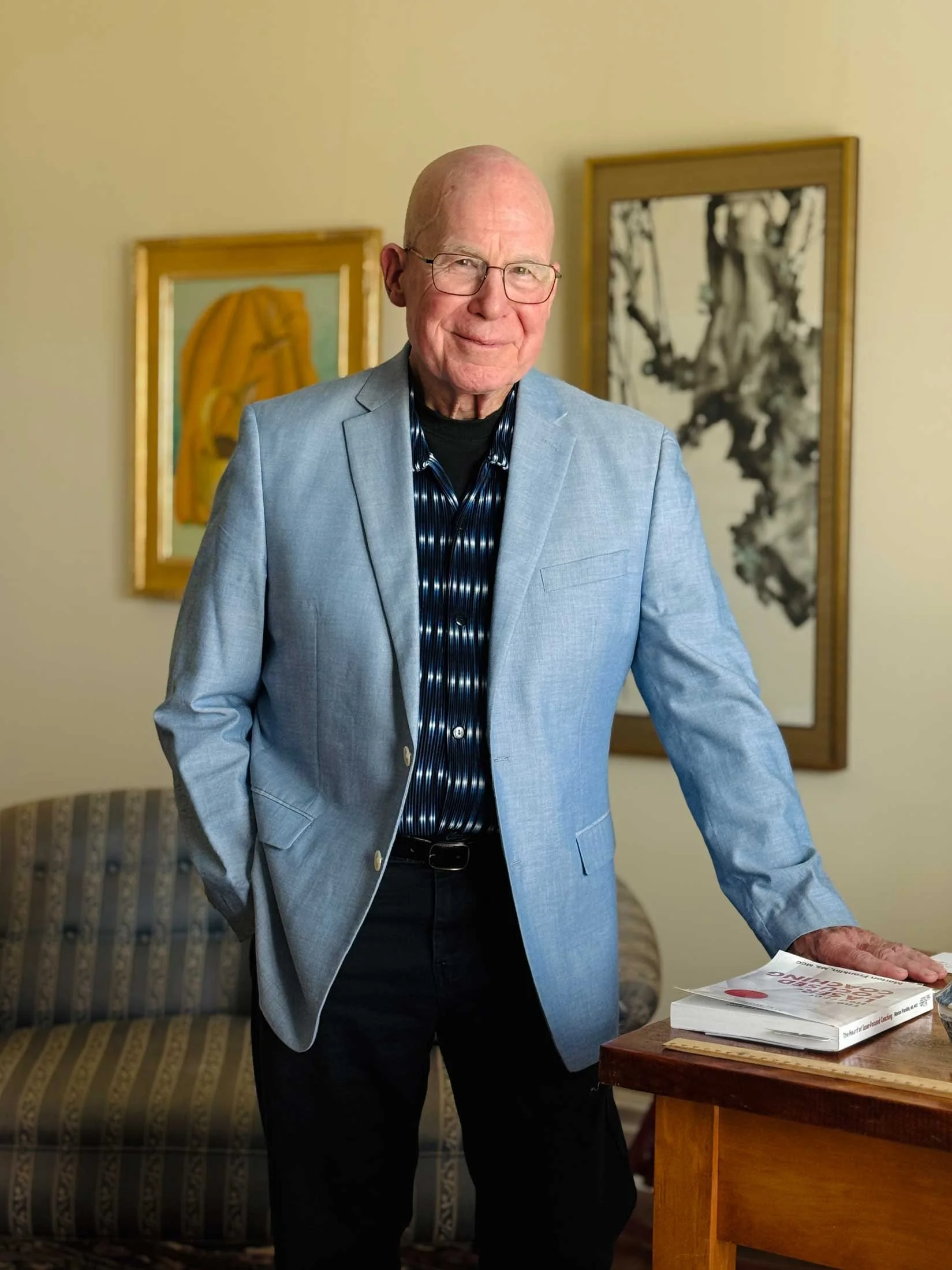 An elderly man with glasses and a light blue blazer standing in a room with artwork on the walls.