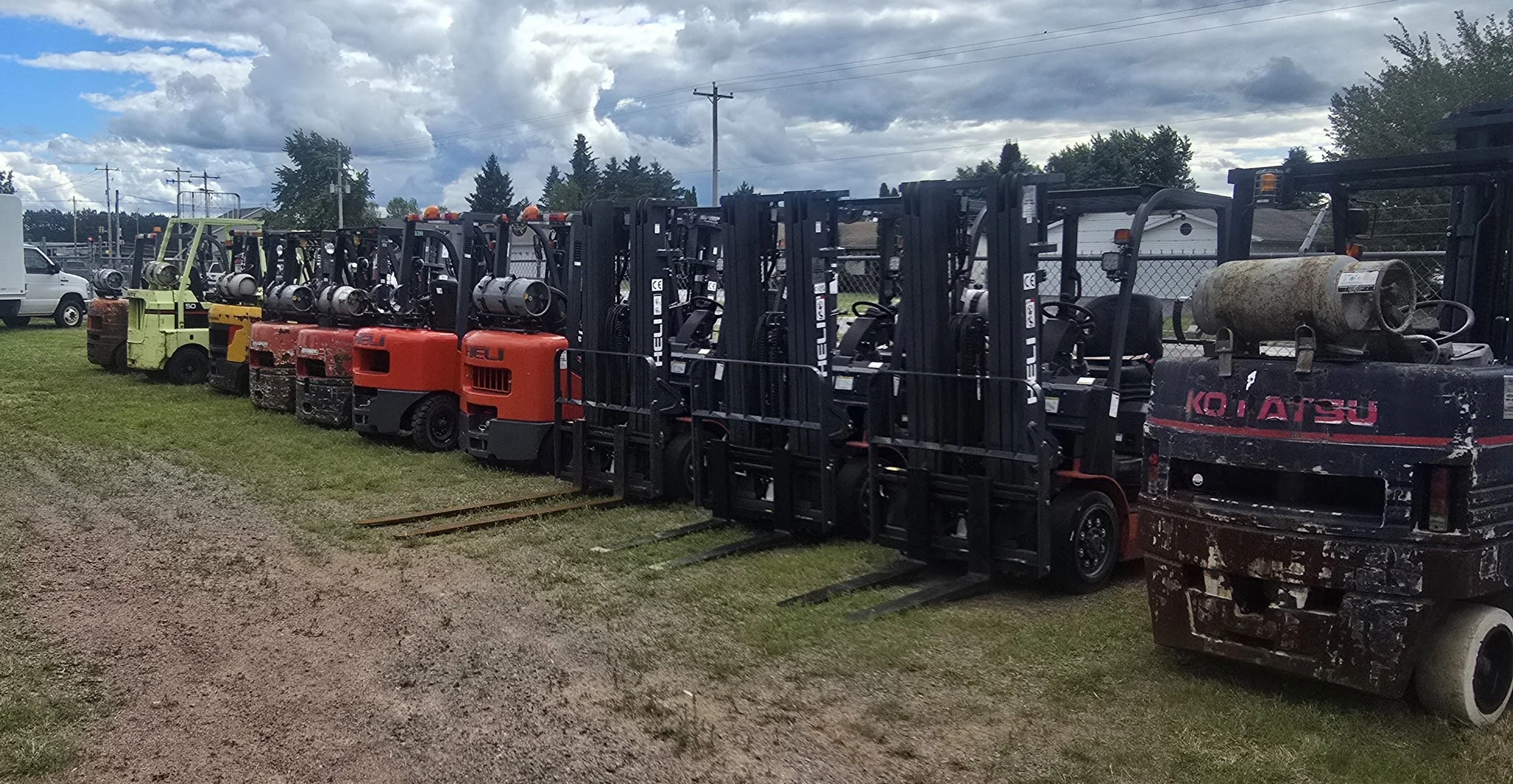 Line of several forklifts parked on grass under cloudy sky, some with gas tanks and signs.