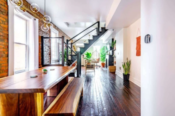 Bright, modern interior of a home with hardwood floors, a staircase with black railing, large windows, and various potted plants.