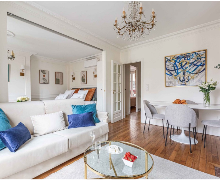 Living room with white walls, a chandelier, a white sofa with blue and white pillows, glass coffee table with strawberries, champagne glasses, bedroom with bed and artwork, dining area with white table, gray chairs, and a large colorful painting.