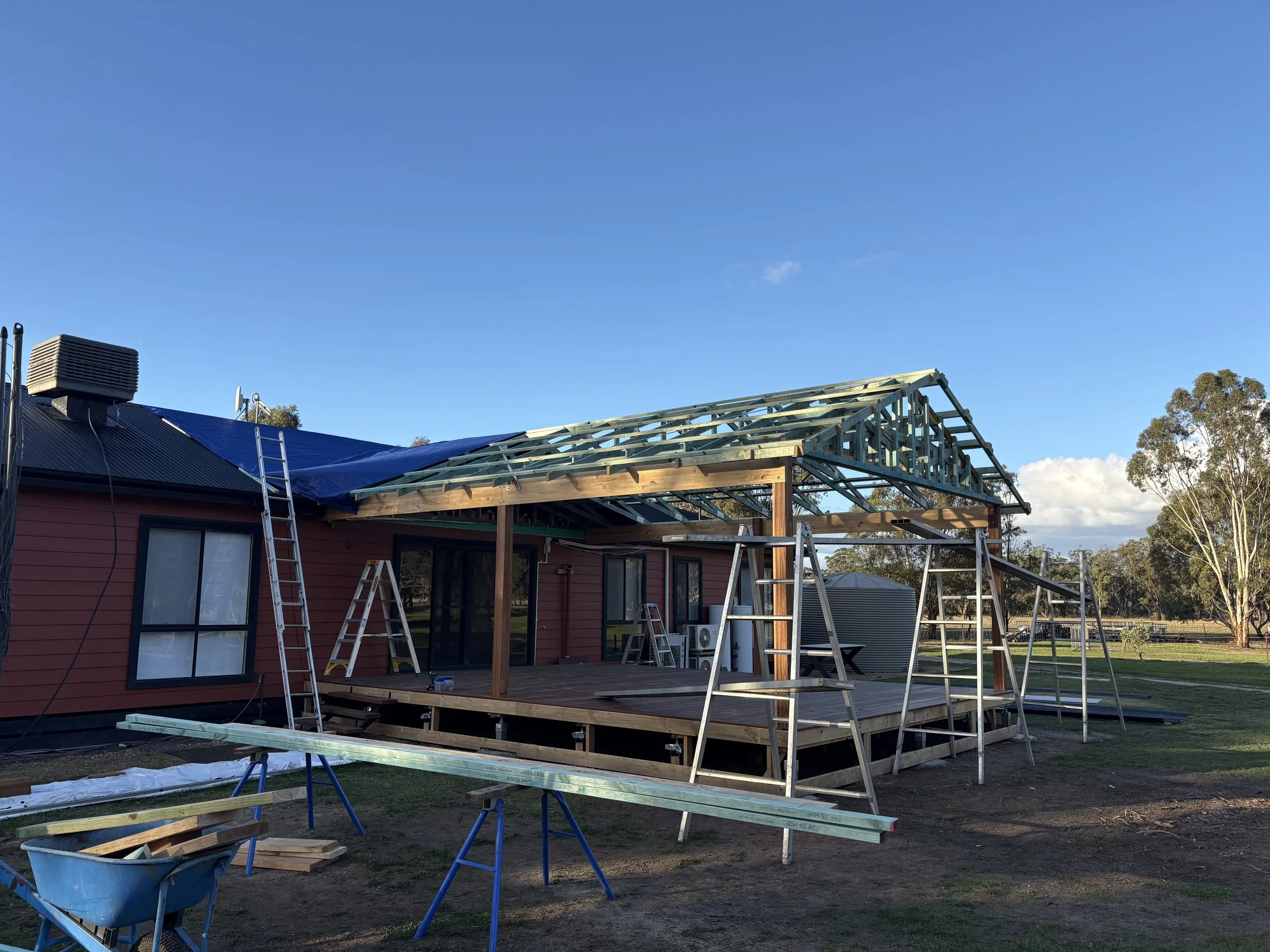 A house under construction with a metal roof, a partially built wooden extension, ladders, and construction tools, set against a blue sky and surrounded by trees.