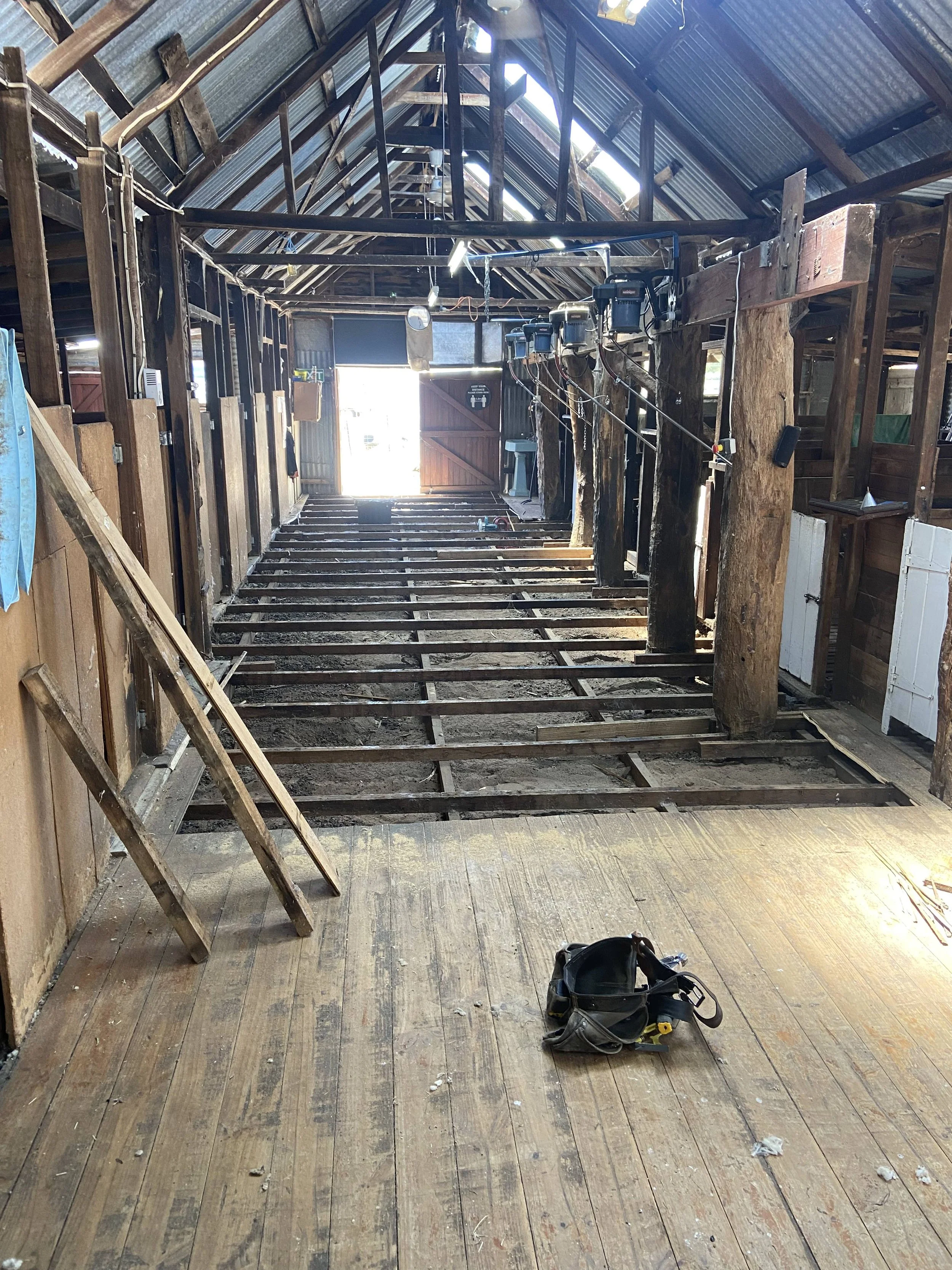 Interior of a barn or shed under renovation, with exposed wooden floor joists, tools, and construction equipment visible, including a ladder and a tool bag on the floor.