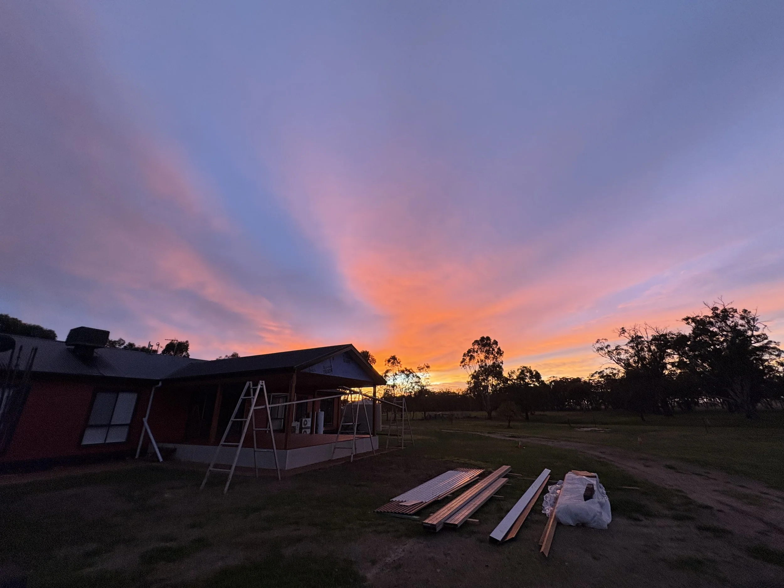 A sunset sky with shades of orange, pink, purple, and blue, with some trees and a building with a metal roof in the foreground, and construction materials lying on the ground.