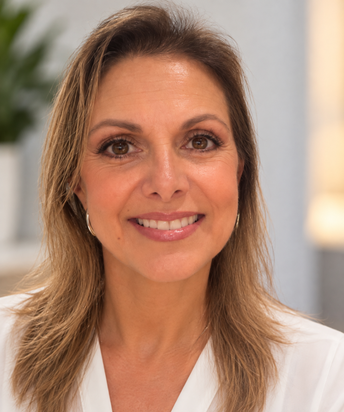 A smiling woman with shoulder-length light brown hair and brown eyes, wearing hoop earrings and a white top, in a well-lit indoor setting.