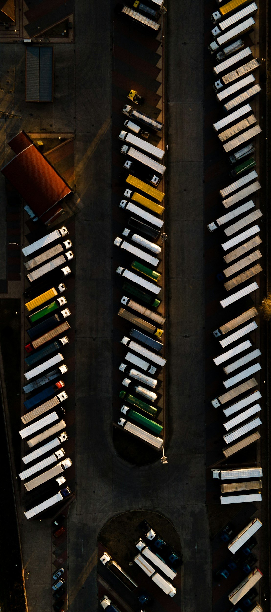 Overhead aerial view of a parking lot filled with semi-trucks parked in organized rows, with some smaller vehicles and buildings visible around the lot.