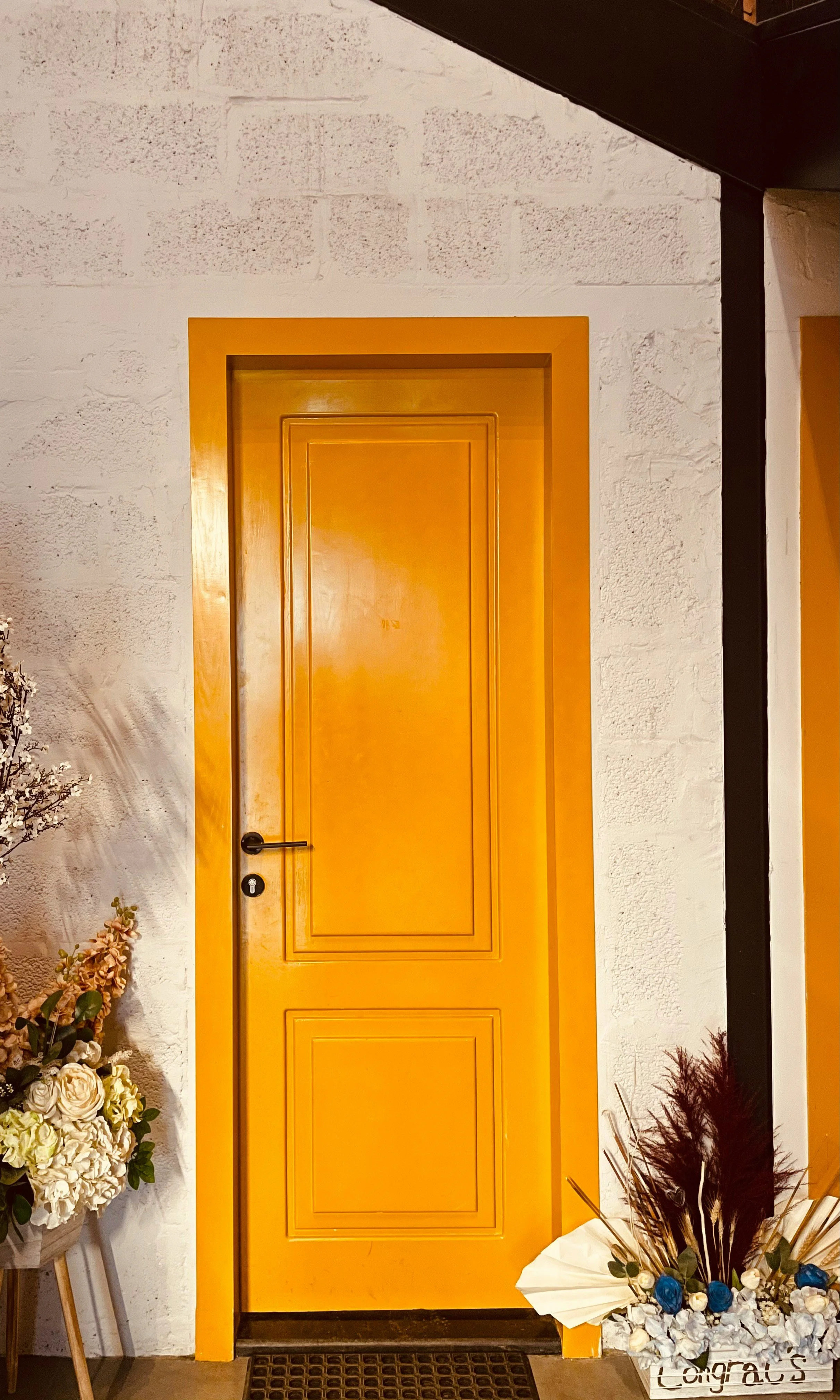 A yellow door with a rectangular inset design, surrounded by a white brick wall. To the left of the door, there is a flower arrangement with white and pale pink flowers in a wooden stand. On the right, there is a large white sign with the word 'Congrats' decorated with blue and white ornaments, alongside a bundle of dark decorative grasses.