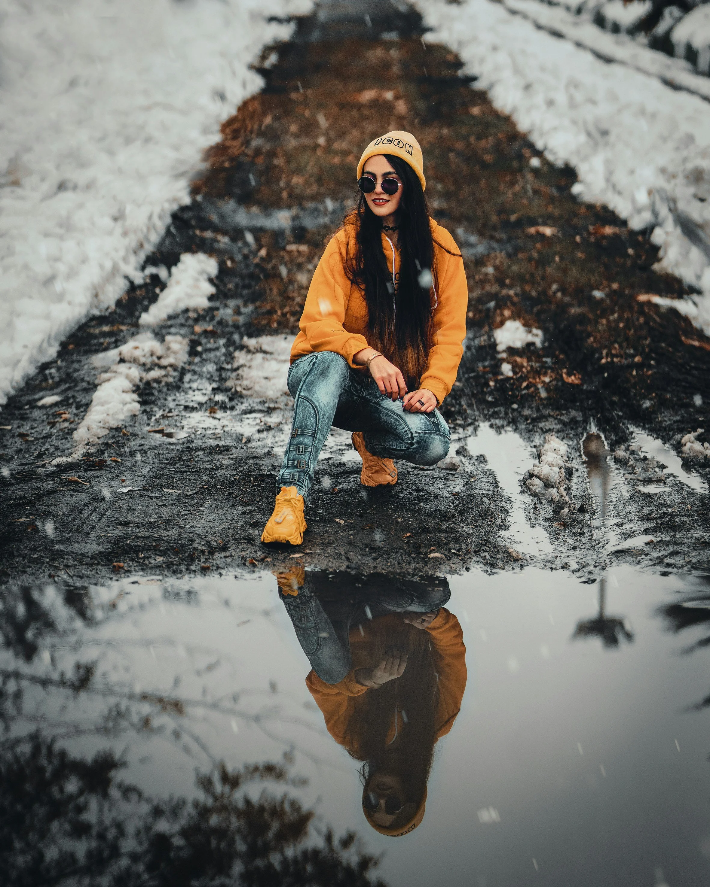 A woman with long dark hair wearing sunglasses, a yellow beanie, and an orange jacket kneels on the ground in a snowy outdoor area, with her reflection visible in a puddle of water.