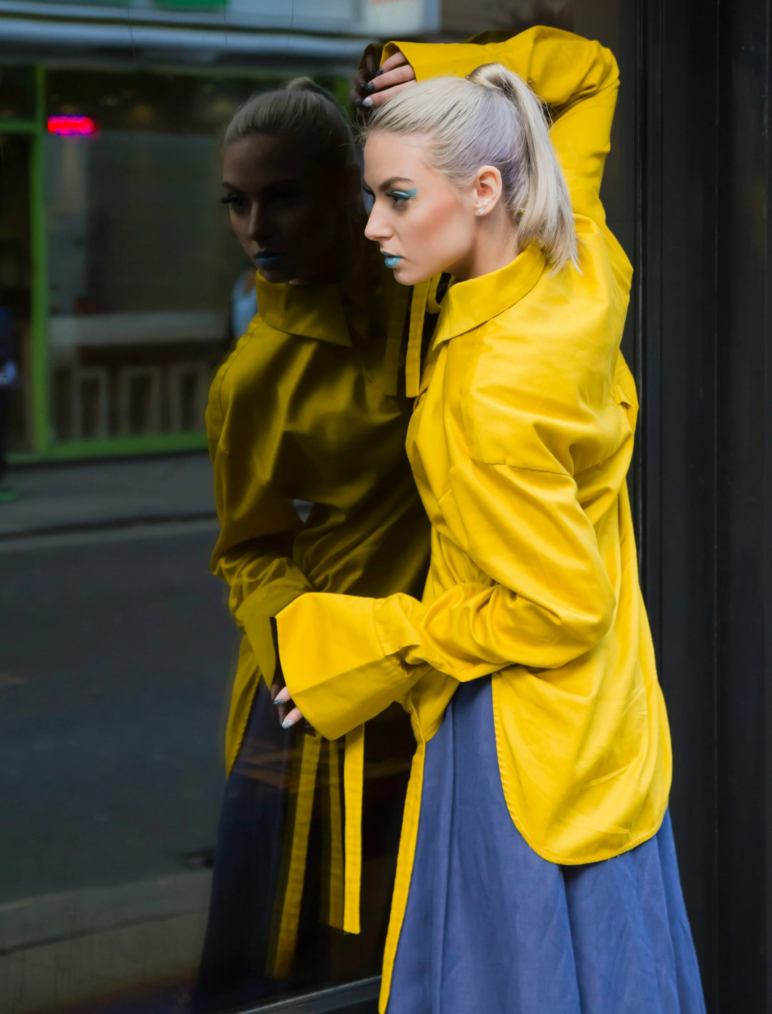 A woman with blonde hair wearing a yellow jacket and blue skirt, standing in front of a mirror in an elevator, with her reflection visible.